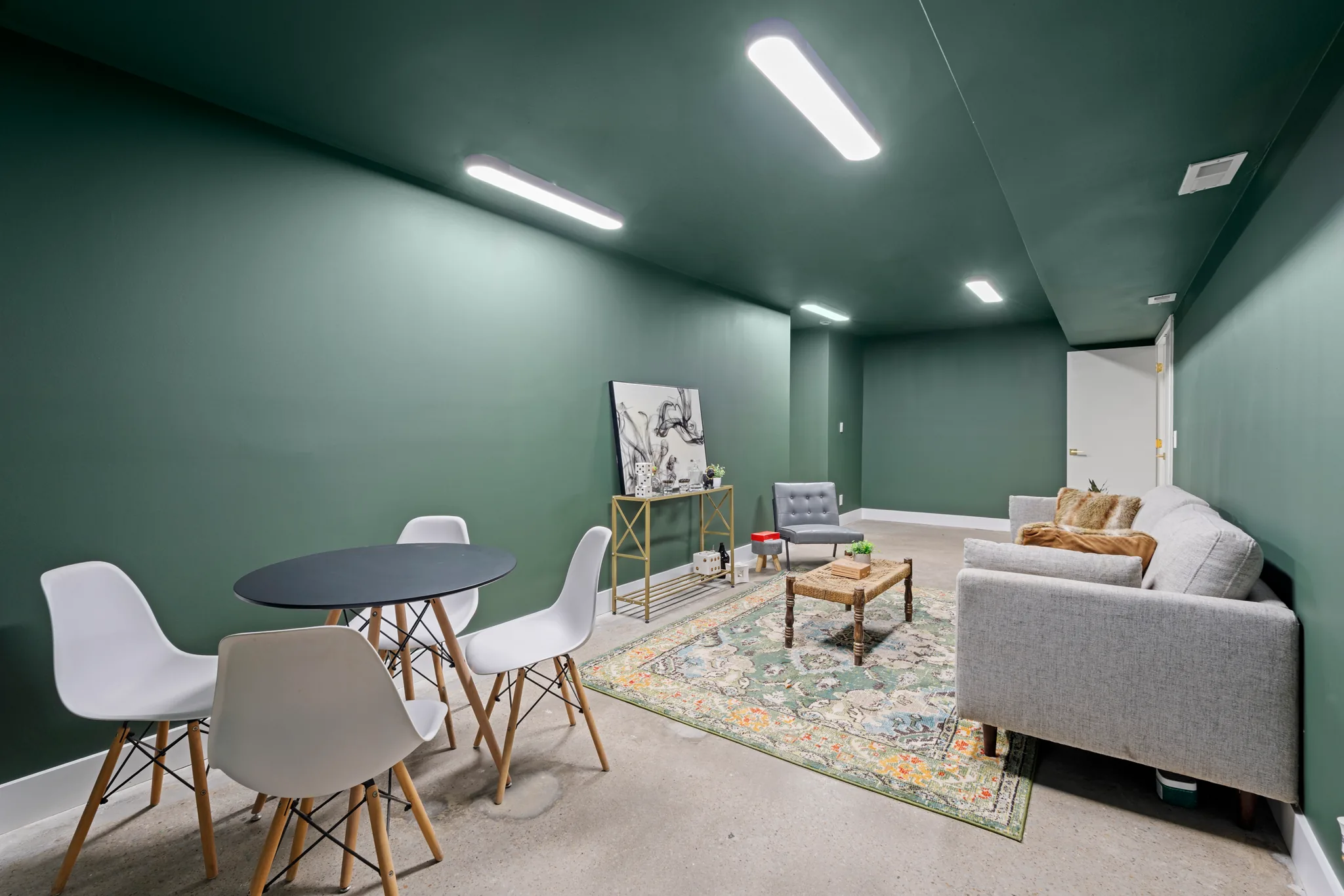 Finished basement room with dark green walls and ceiling, polished concrete floor, white dining chairs, gray sofa and patterned rug