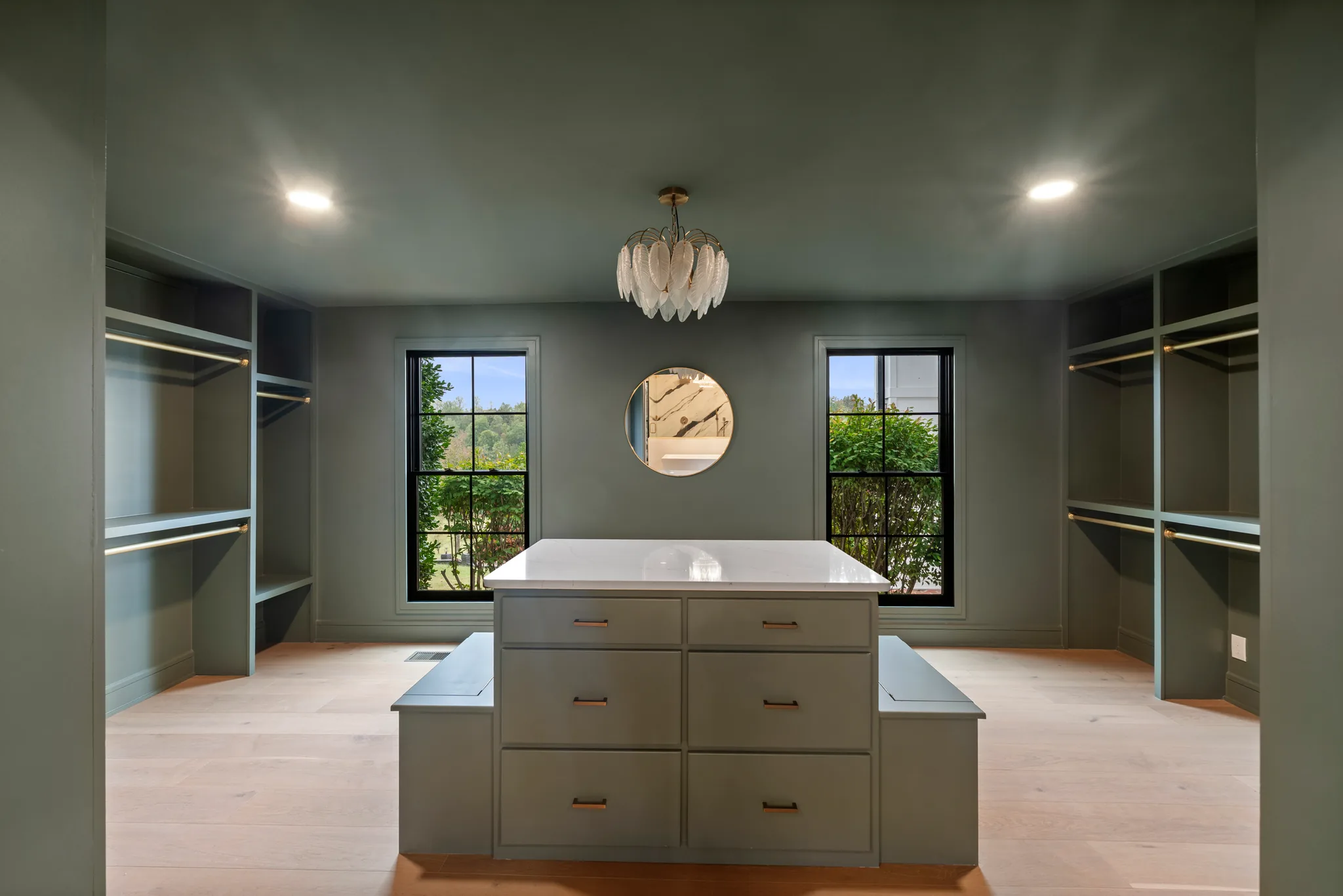 Walk-in closet with green painted walls and built-in shelving, a six-drawer island with white stone top, round mirror and two black grid windows