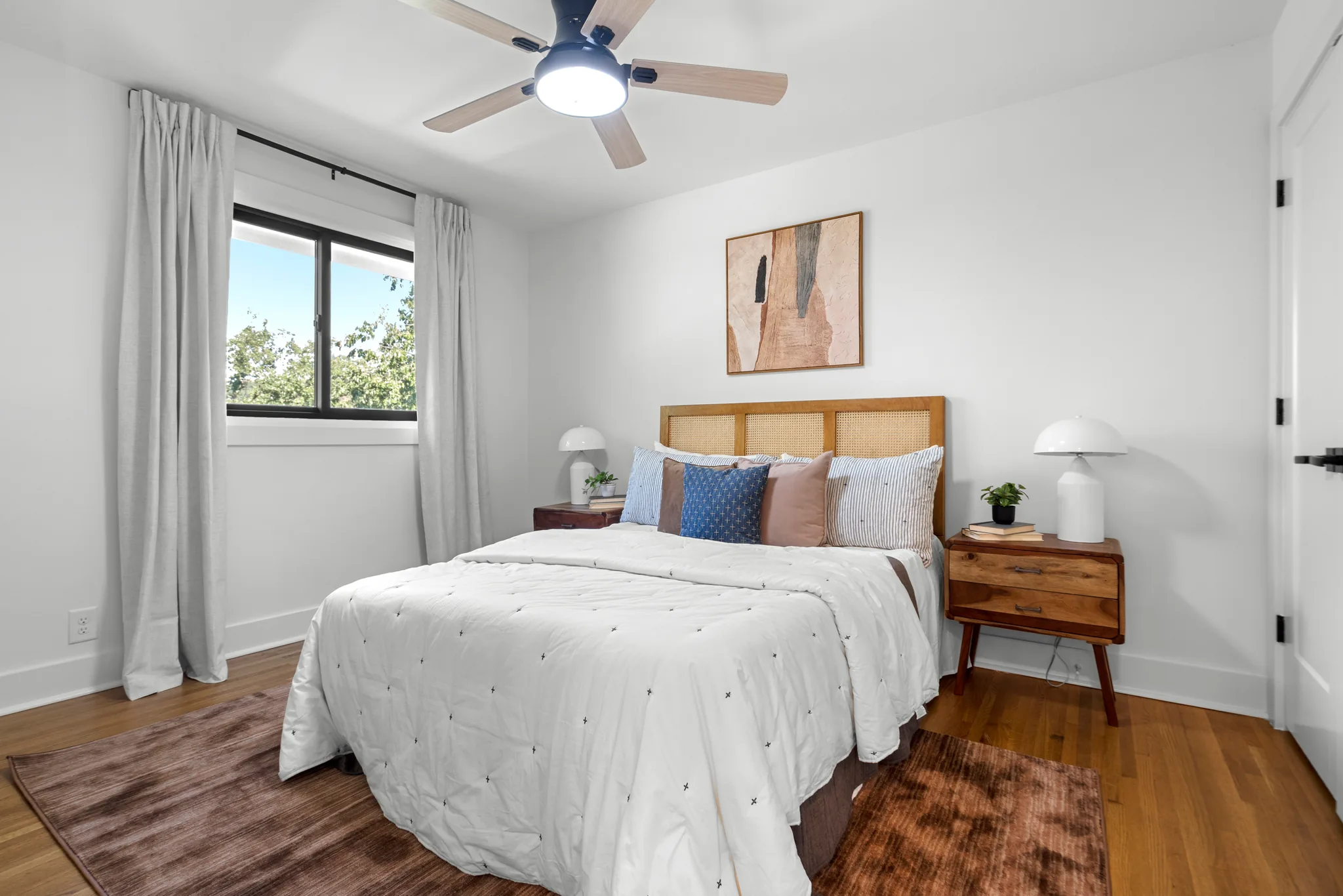 Bedroom with white walls, oak floors, a cane headboard bed, wood nightstands, black-framed window with linen curtains and a ceiling fan