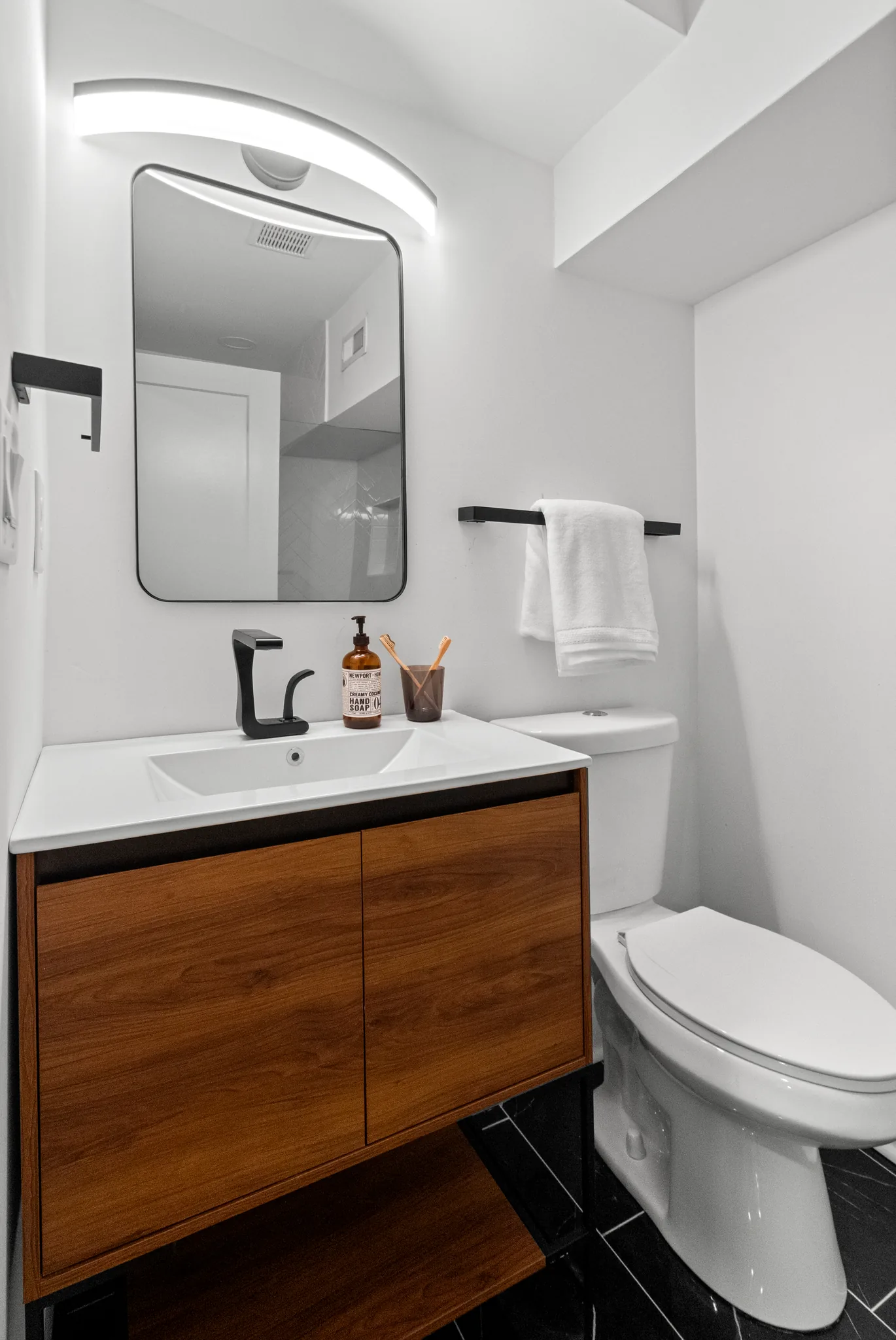 Small bathroom with a wall-mounted walnut vanity, white integrated sink, black faucet, rounded rectangular mirror, black towel bar and dark tile floor
