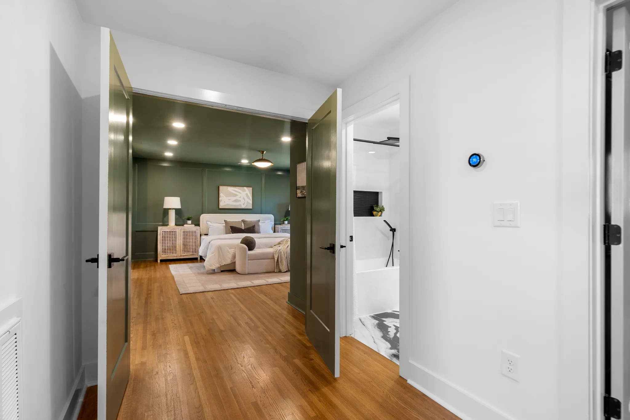 Hallway with red oak floors and green painted double doors opening into a green bedroom, with a white bathroom to the right