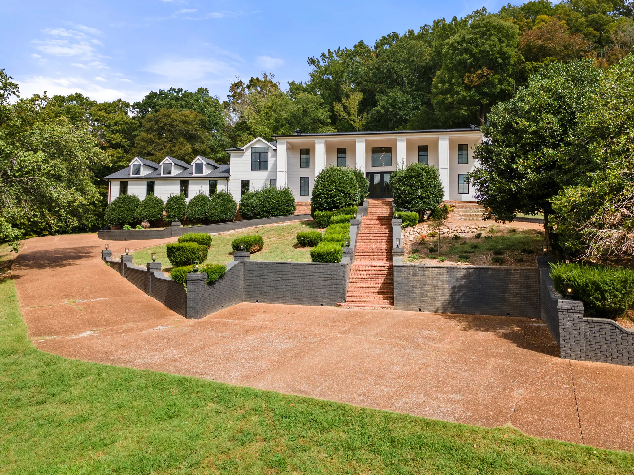 White two-story home on a wooded hillside with columned entry, dark gray block retaining walls and a brick stairway rising from a wide exposed-aggregate drive