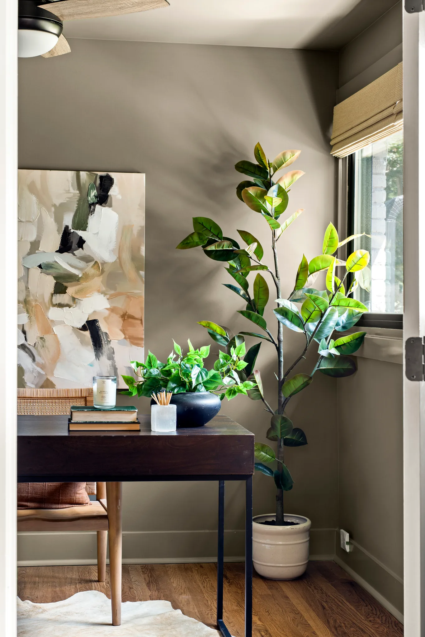 Corner home office with dark wood desk, abstract canvas art, potted plant and window with woven shade above hardwood floors