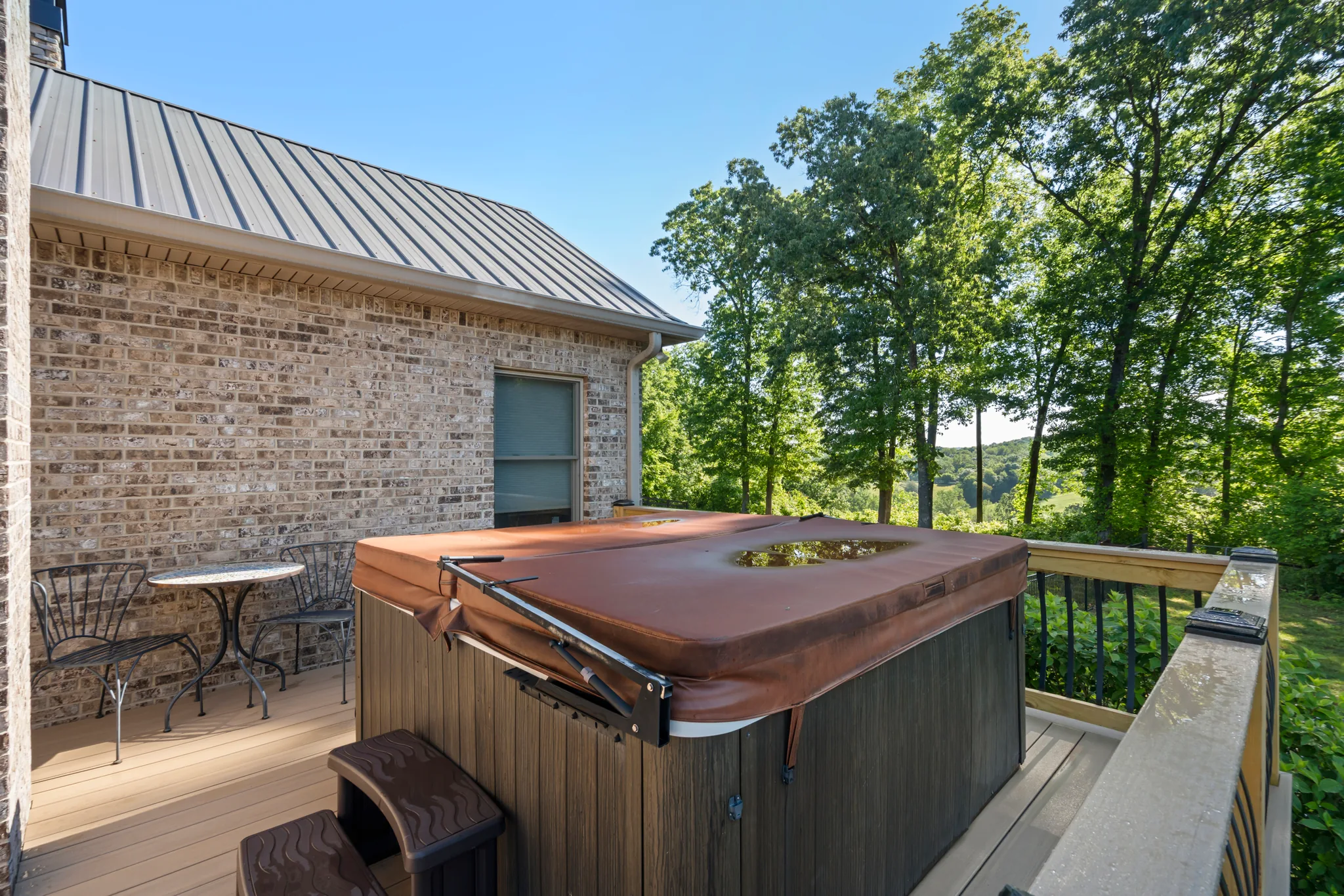 Covered hot tub with a brown cover on a tan composite deck beside a brick wall, with wood railing and wooded valley view