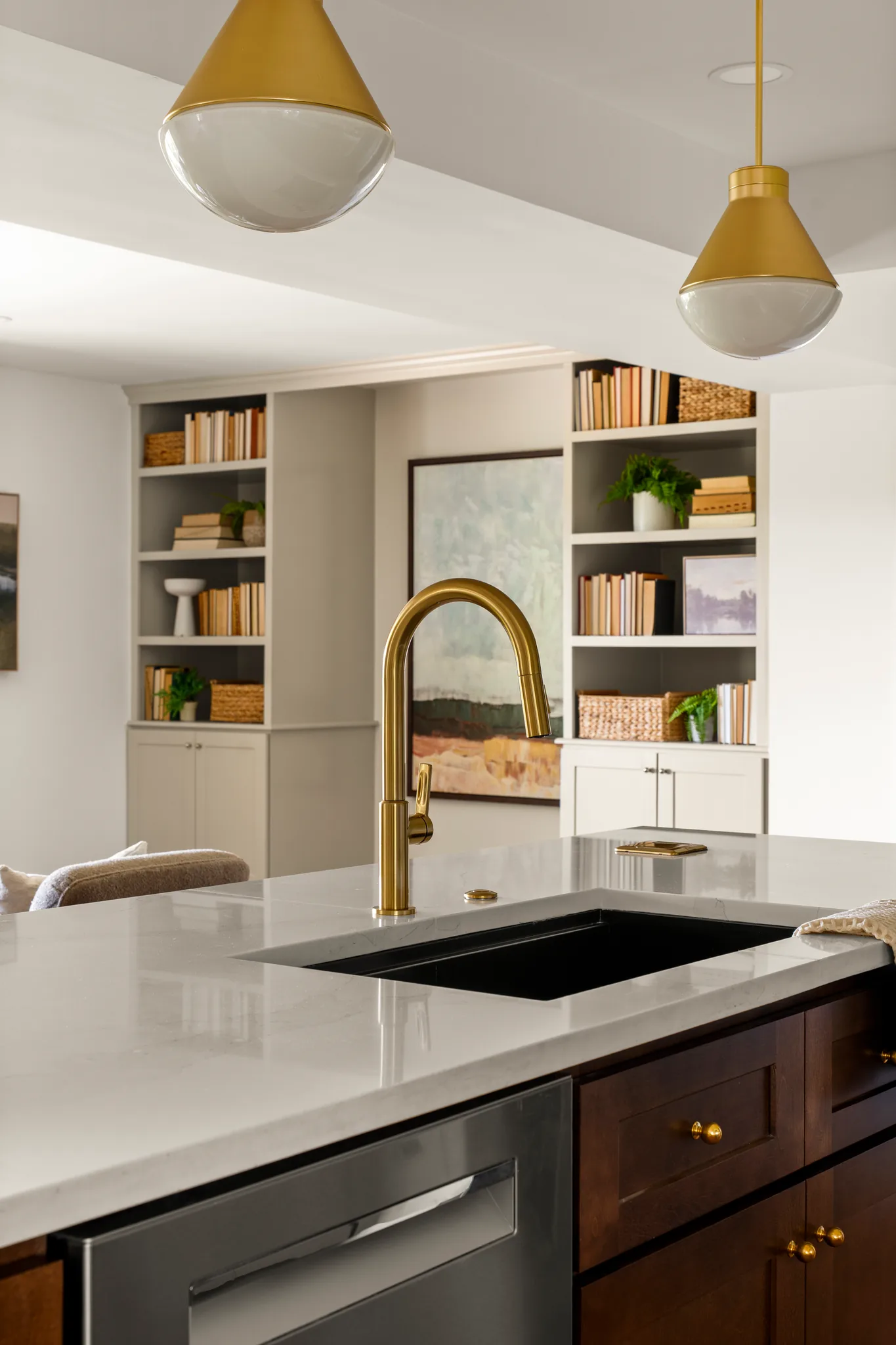 Kitchen island with white quartz counter, black undermount sink, brass gooseneck faucet, dark wood cabinets and brass pendants overhead