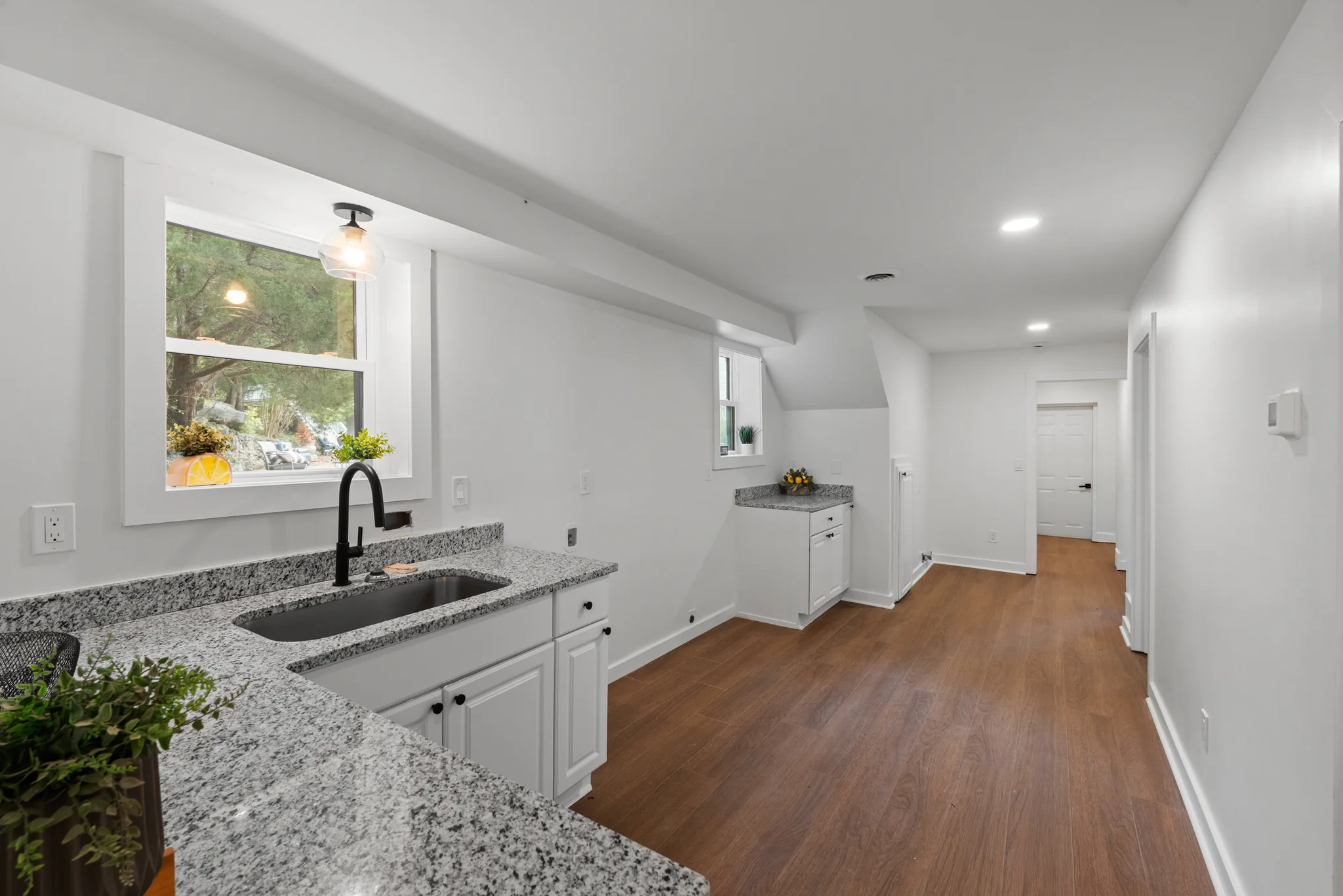 Upper-level kitchenette with speckled granite counters, white shaker cabinets, black faucet and undermount sink, opening to a hallway with wood-look flooring