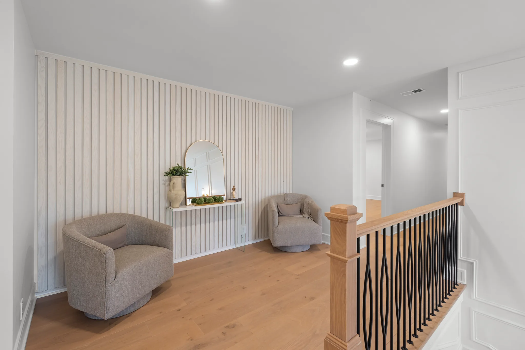 Upstairs landing sitting area with a white vertical slat accent wall, two gray swivel chairs, console with arched mirror and oak stair railing