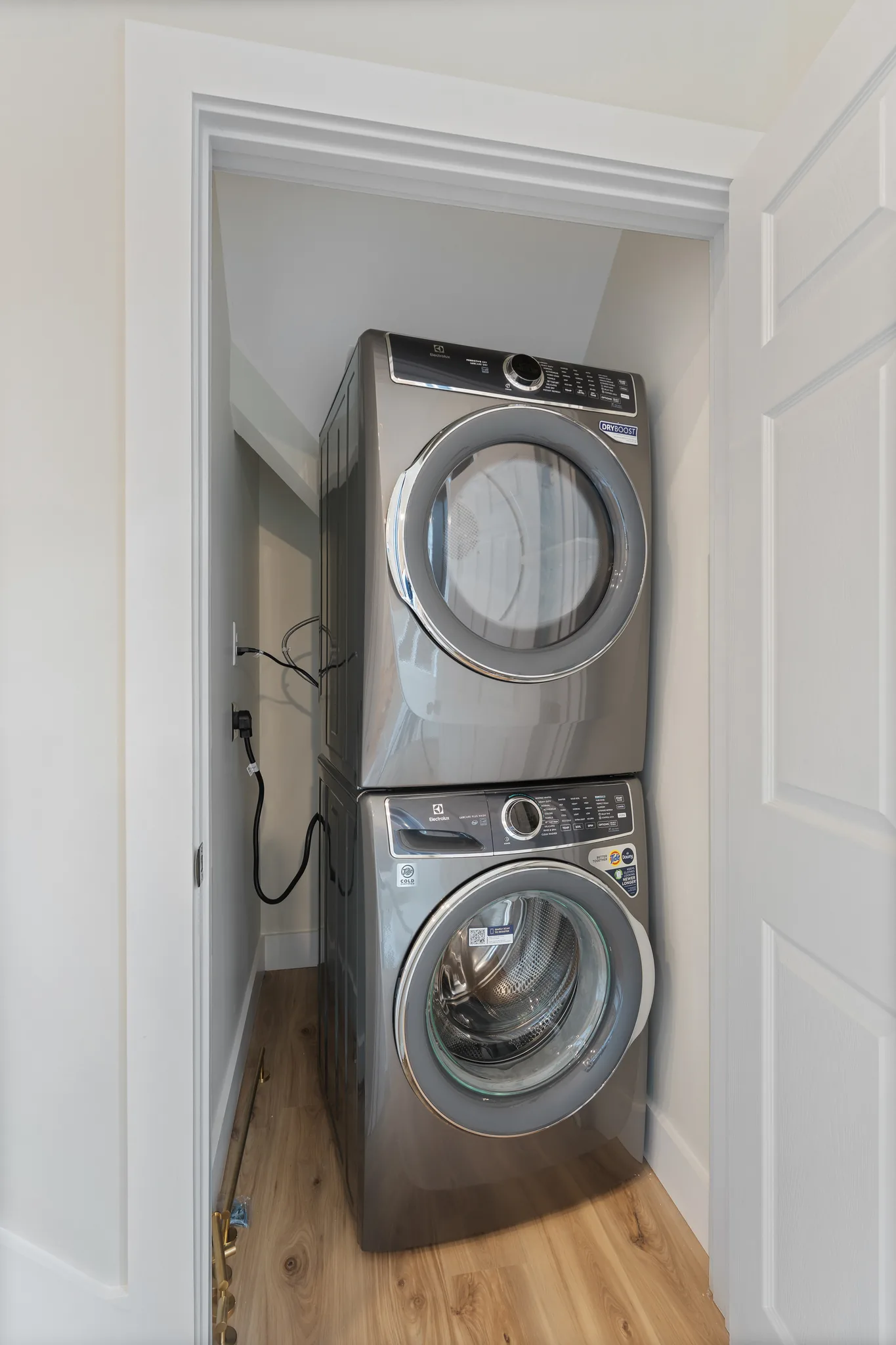Stacked front-load washer and dryer in a laundry closet with white trim, gray walls and light wood plank flooring