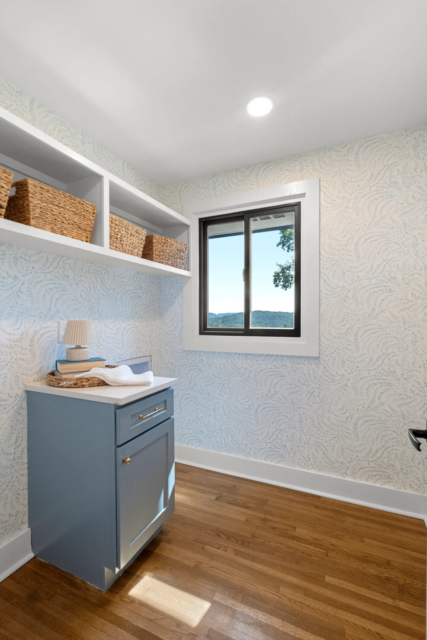 Small utility room with light blue patterned wallpaper, blue-gray base cabinet, white open shelves holding woven baskets, and oak floors