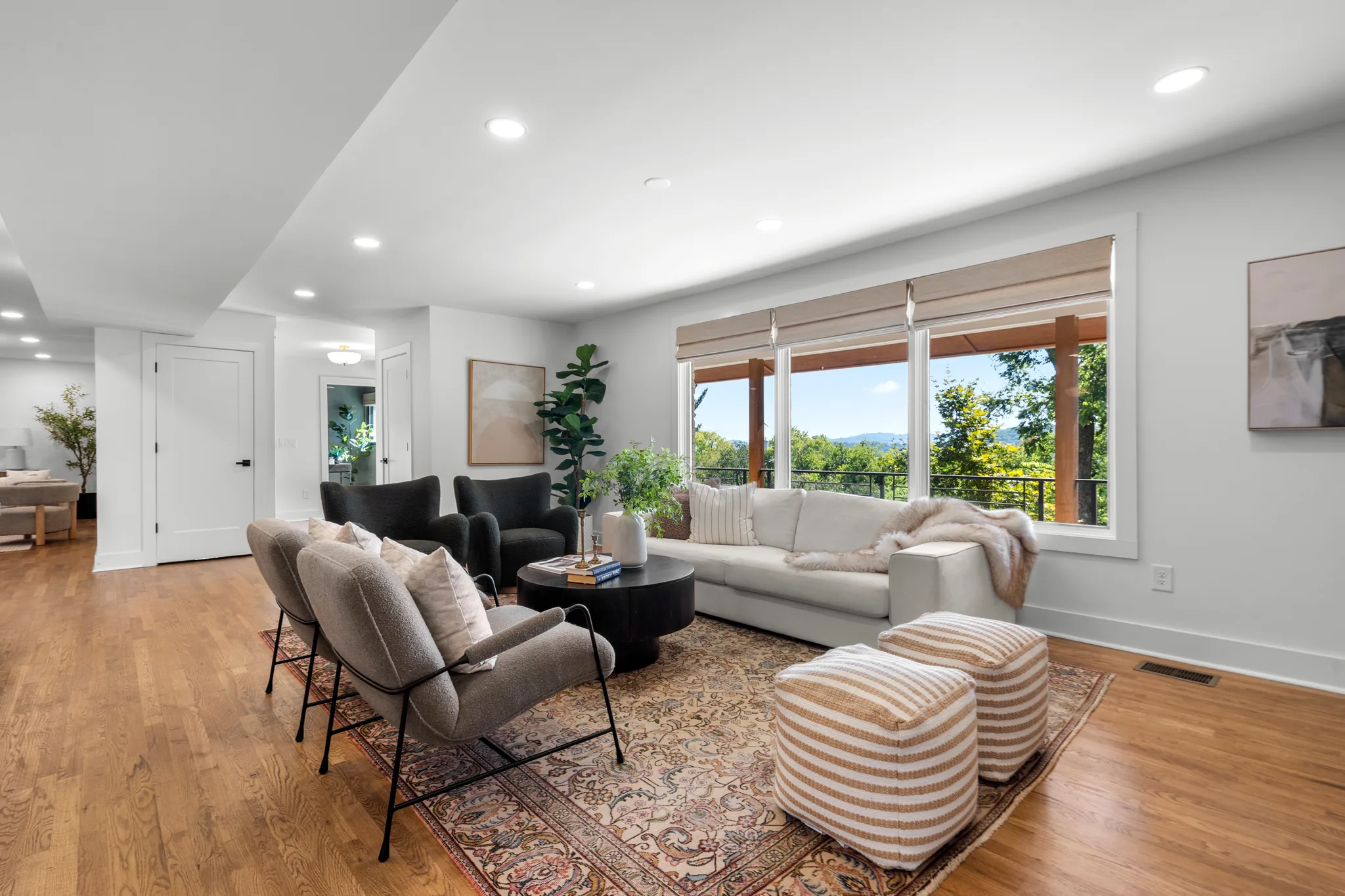 Open living area with oak floors, white walls, recessed lighting, and large wood-framed windows looking out to a deck and wooded hillside