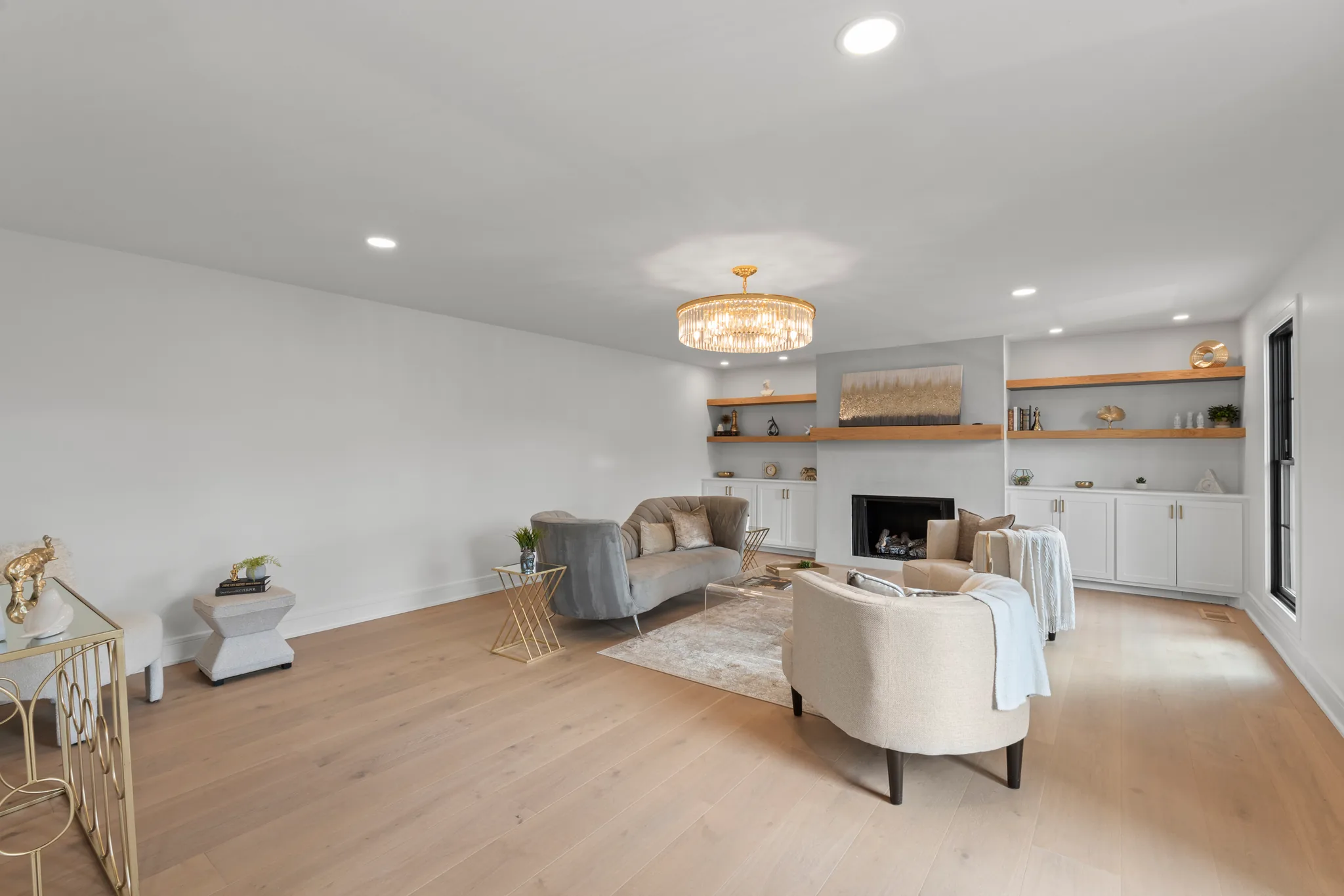 Wide living room with white walls, light oak plank floors, a gray plaster fireplace flanked by white cabinets, wood shelves and a tiered brass chandelier