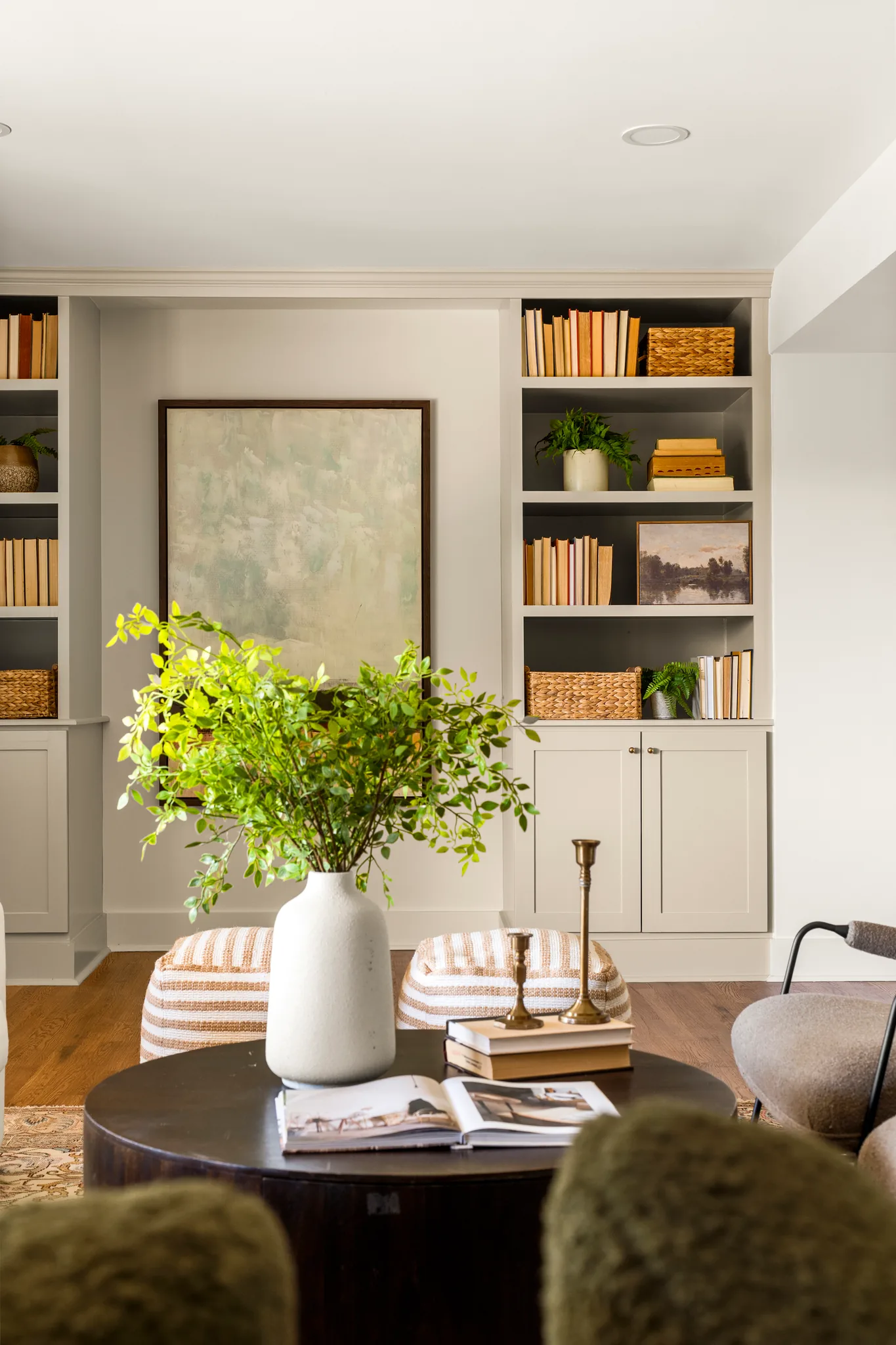 Living room with painted built-in shelving and shaker base cabinets flanking a framed painting, above hardwood floors with a round dark coffee table