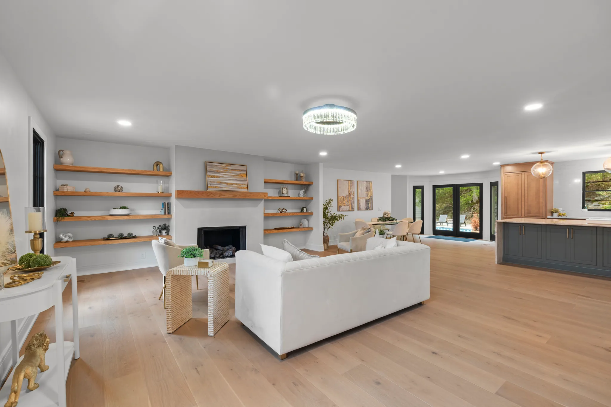 Open living room with white plaster fireplace, oak floating shelves, light wide-plank floors and sightlines to a kitchen with dark cabinets