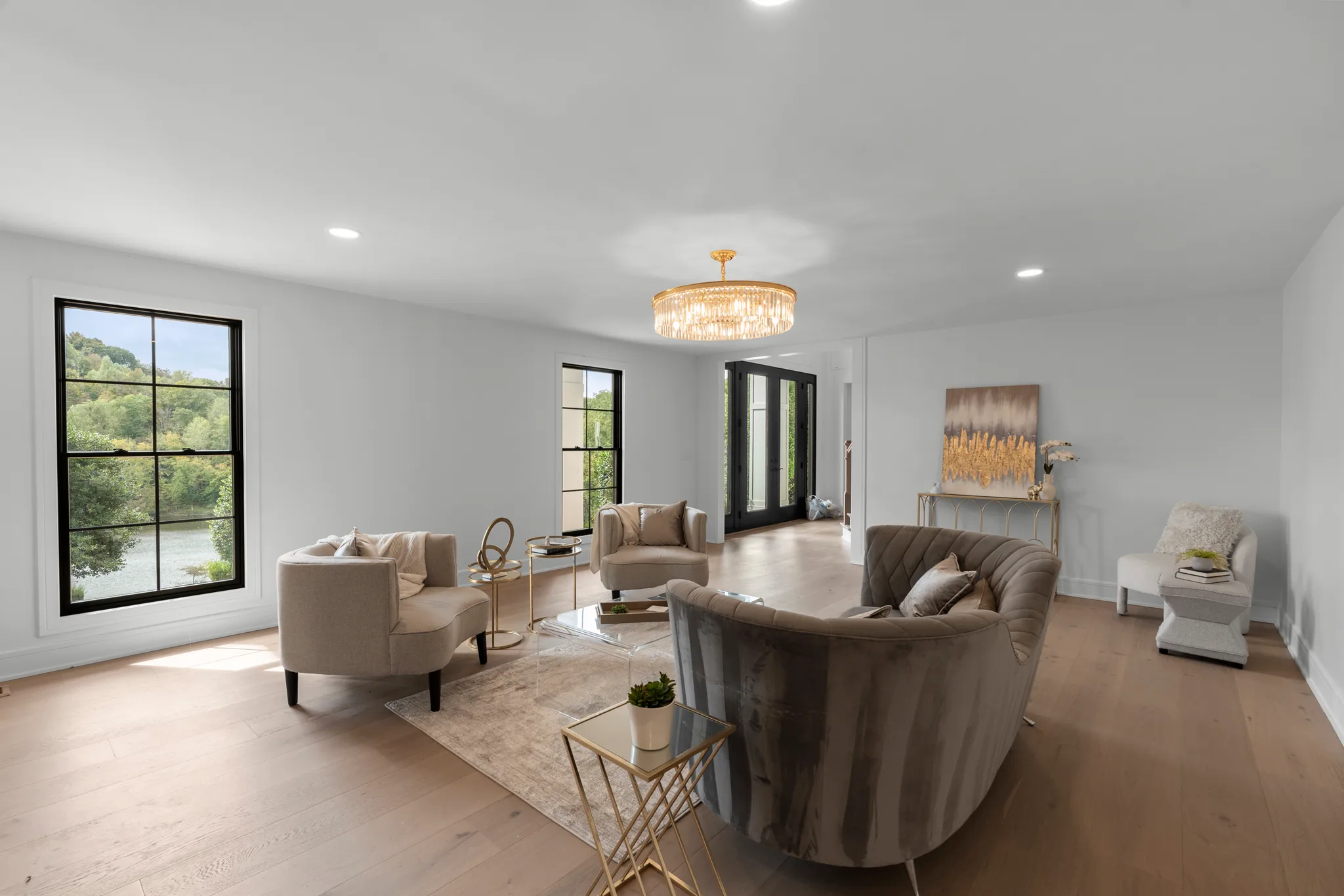 Open living room with white walls, wide oak flooring, black-framed windows overlooking a lake, and a tiered gold chandelier above seating