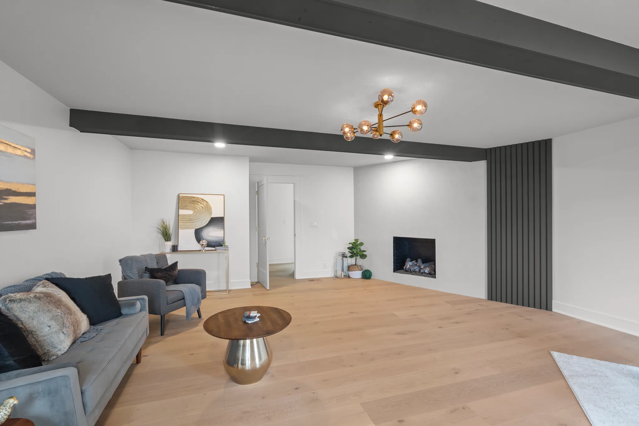 Open living room with white walls, dark painted ceiling beams, plaster fireplace and a black fluted accent panel beside light oak flooring
