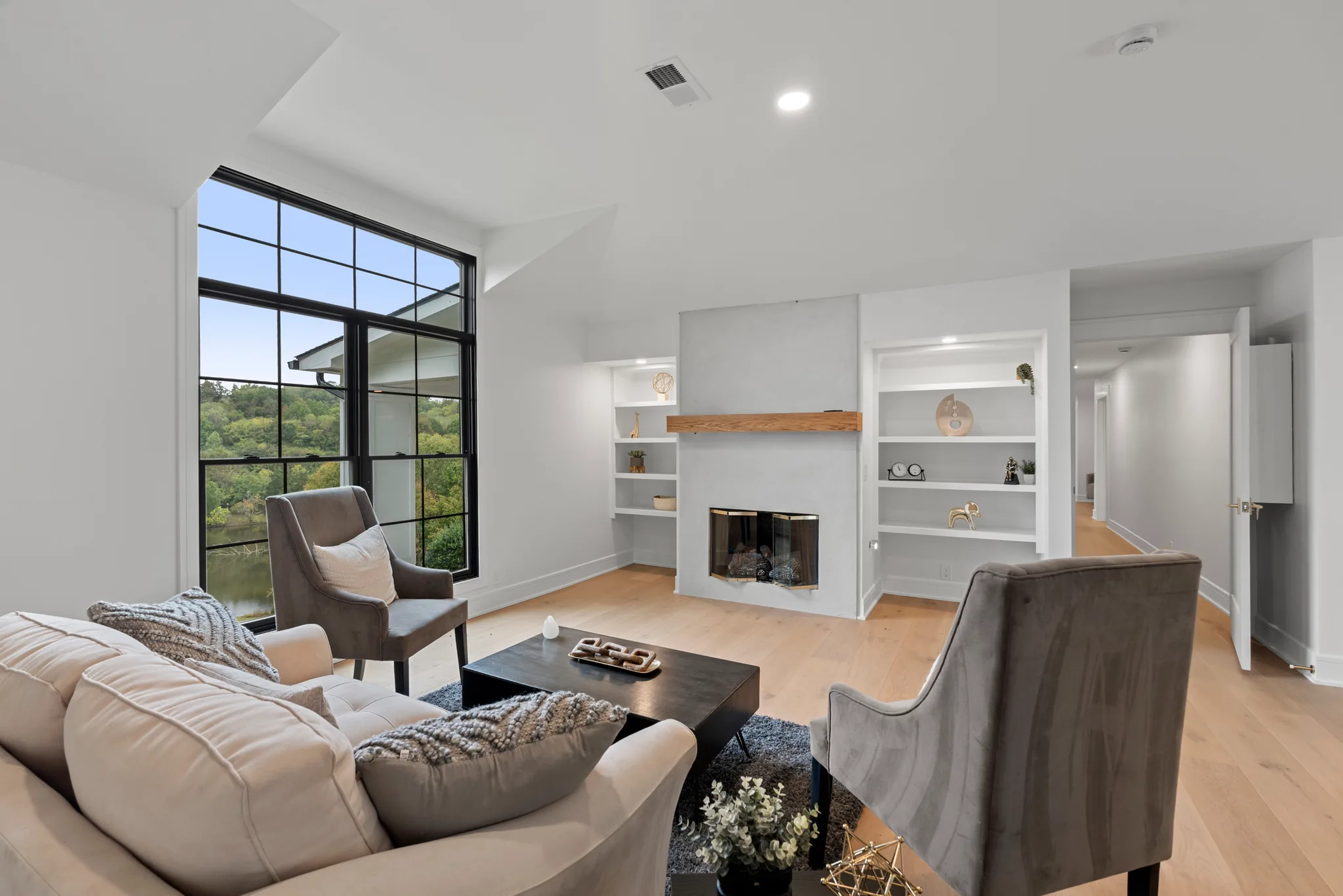 Living room with a light plaster fireplace, wood mantel, white built-in shelves, tall black-framed window and pale oak flooring