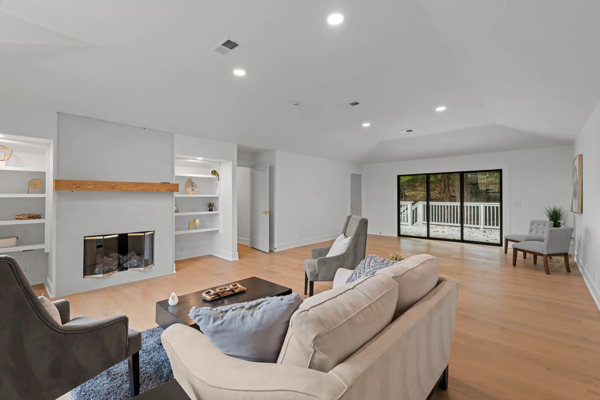 Open living room with a white plaster fireplace, thick wood mantel, flanking built-in shelves, light oak floors and sliding glass doors to a deck