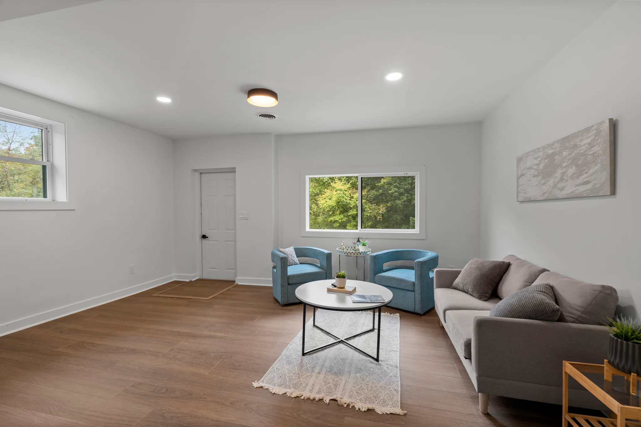 Finished living room with light gray walls, wide plank wood floors, sliding windows to trees, a gray sofa and two blue chairs