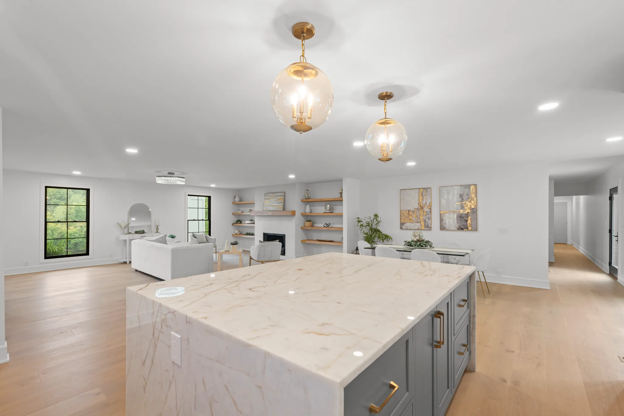 Large kitchen island with veined marble waterfall top and gray cabinets, two glass globe pendants above, open to a living area with fireplace