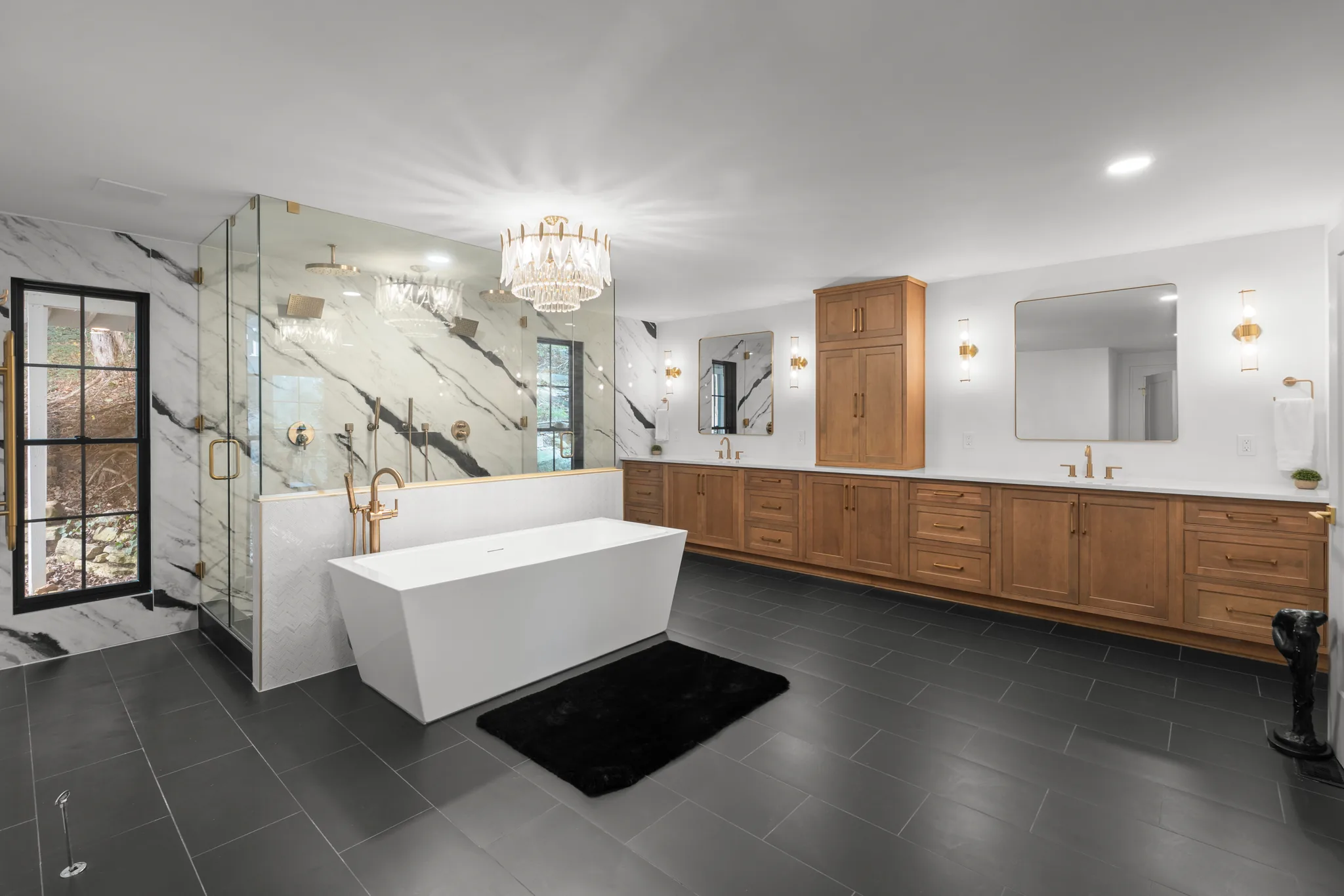 Primary bath with a glass corner shower against veined marble walls, a white freestanding tub, wood vanities and dark gray floor tile