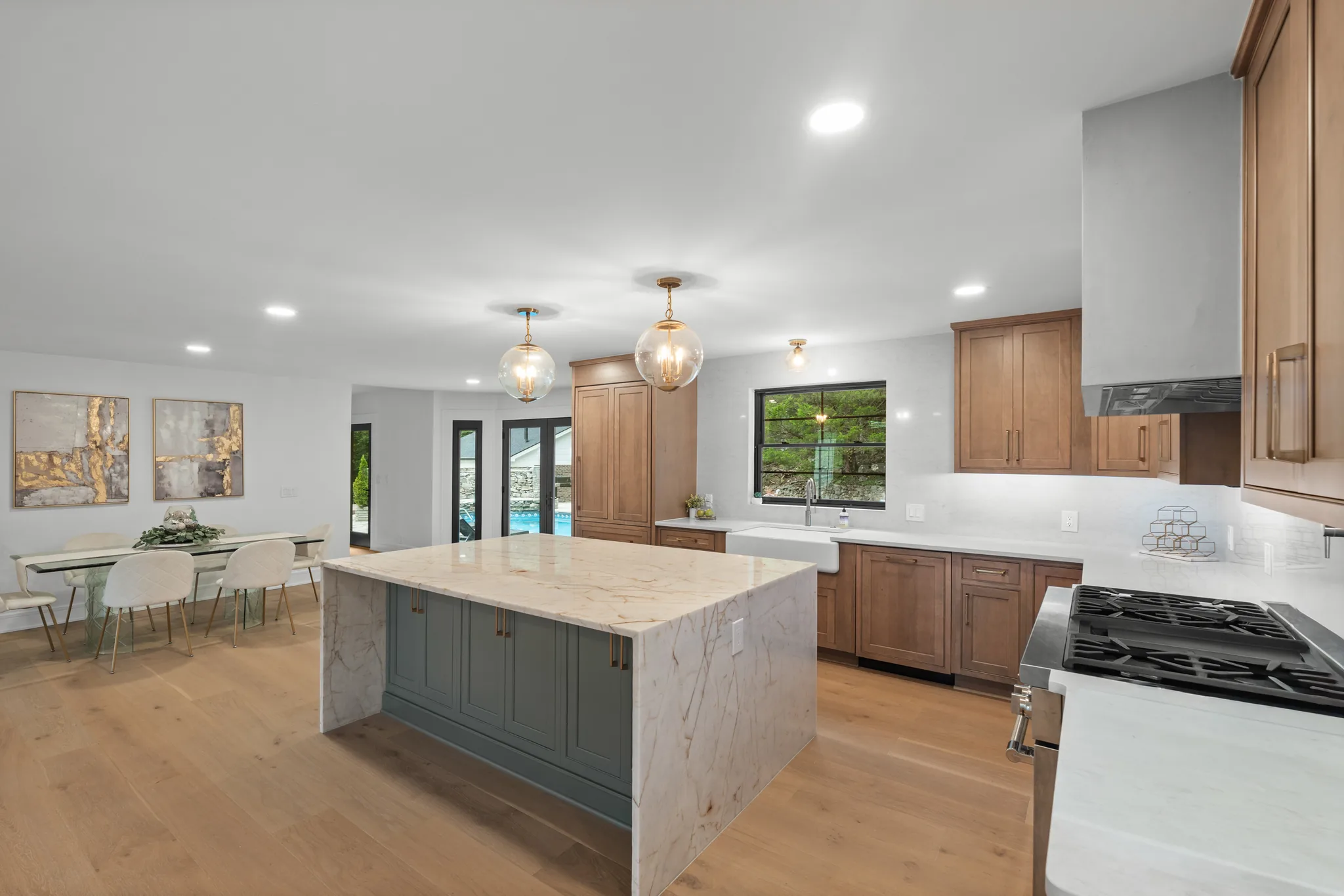 Kitchen with a green painted island under a veined marble waterfall countertop, stained wood cabinets, white farmhouse sink and glass globe pendants