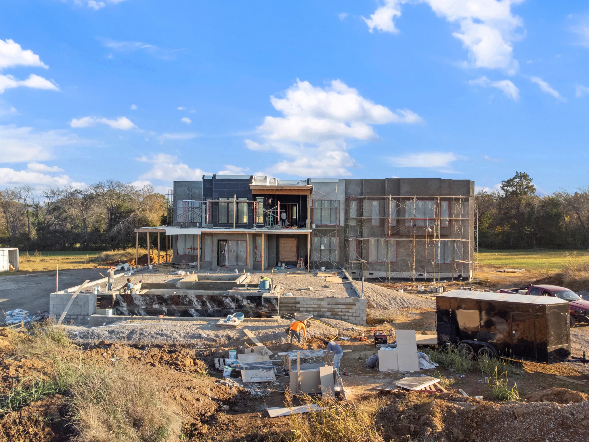 Two-story modern home under construction with scaffolding and sheathed walls, crews working on a concrete pool shell and stone retaining wall below