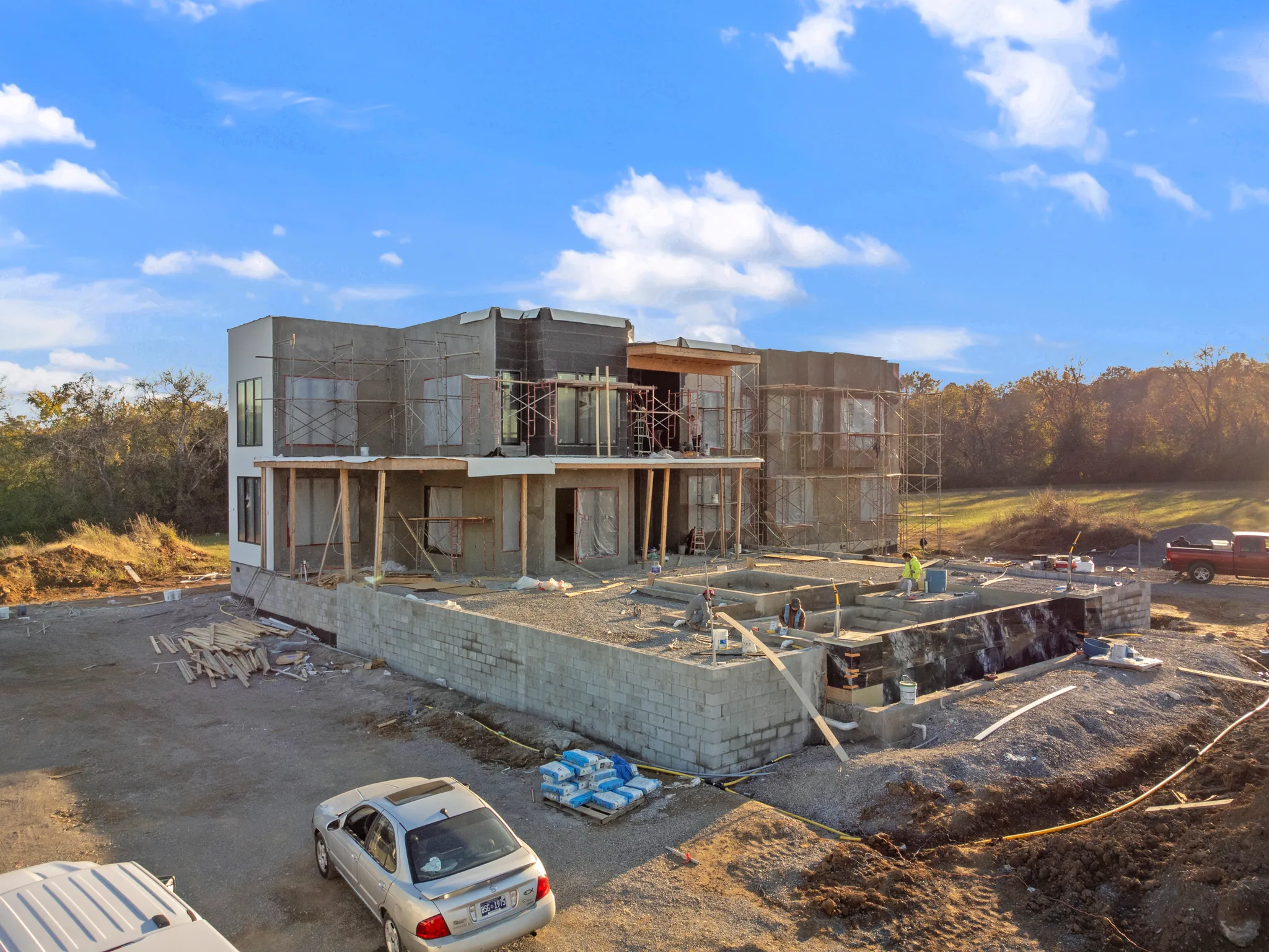 Two-story modern home under construction with scaffolding, sheathed walls and a block foundation for an adjoining pool area, workers on site