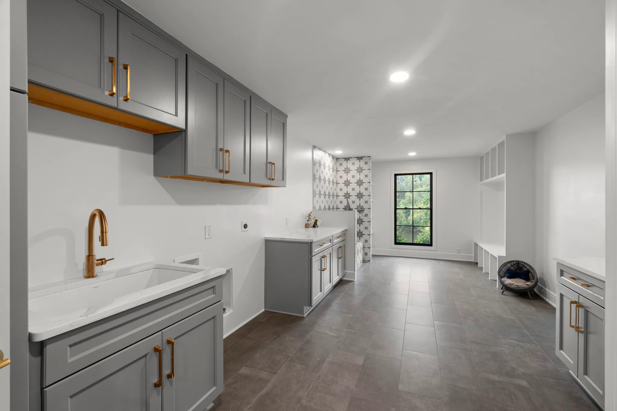 Long mudroom and laundry room with gray cabinets, brass hardware, a quartz utility sink, patterned tile accent wall and built-in bench lockers
