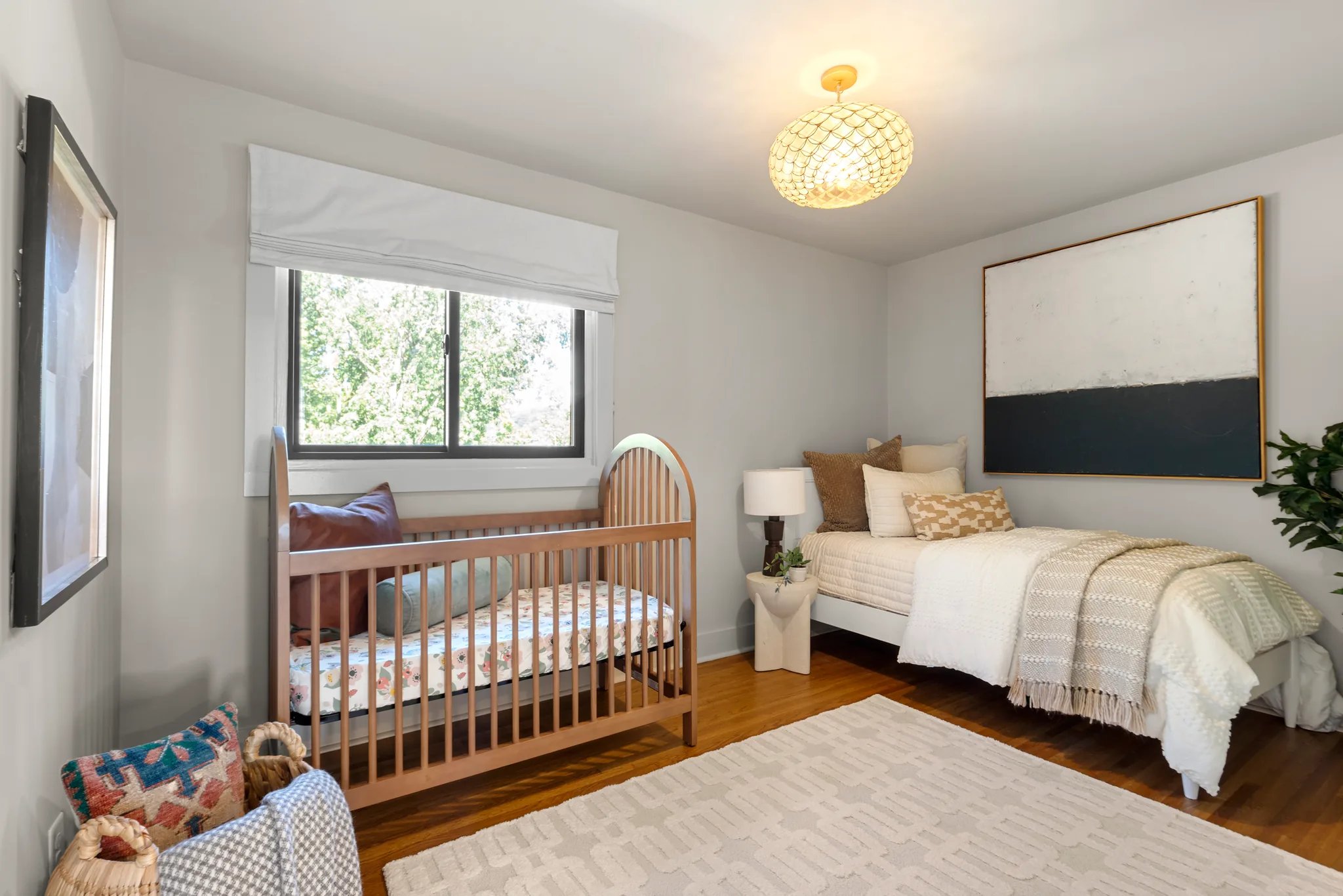 Child's bedroom with a wood crib, twin bed, hardwood floors, light gray walls, a shaded window and a woven ceiling light