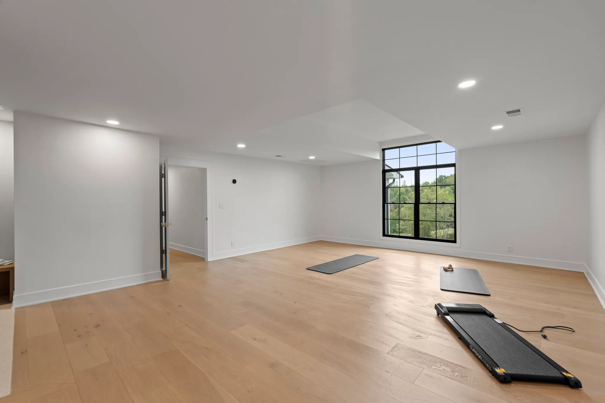 Empty lower level exercise room with light oak plank floors, white walls, recessed lights, a tall black grid window and exercise mats