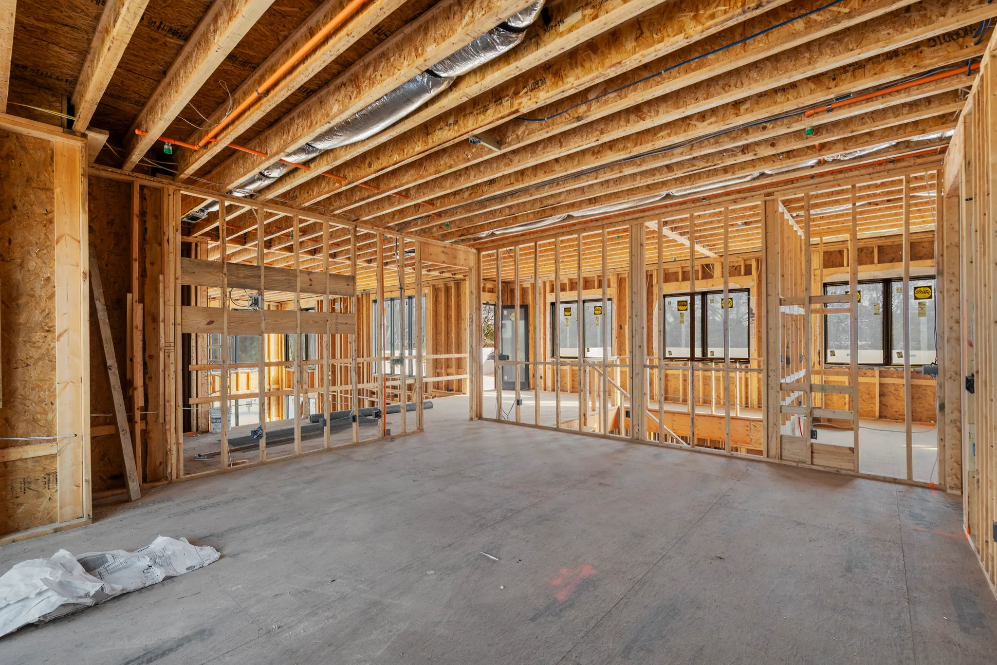 Interior framing under construction with exposed ceiling joists, insulated duct runs and electrical wiring above a bare concrete floor and installed black windows