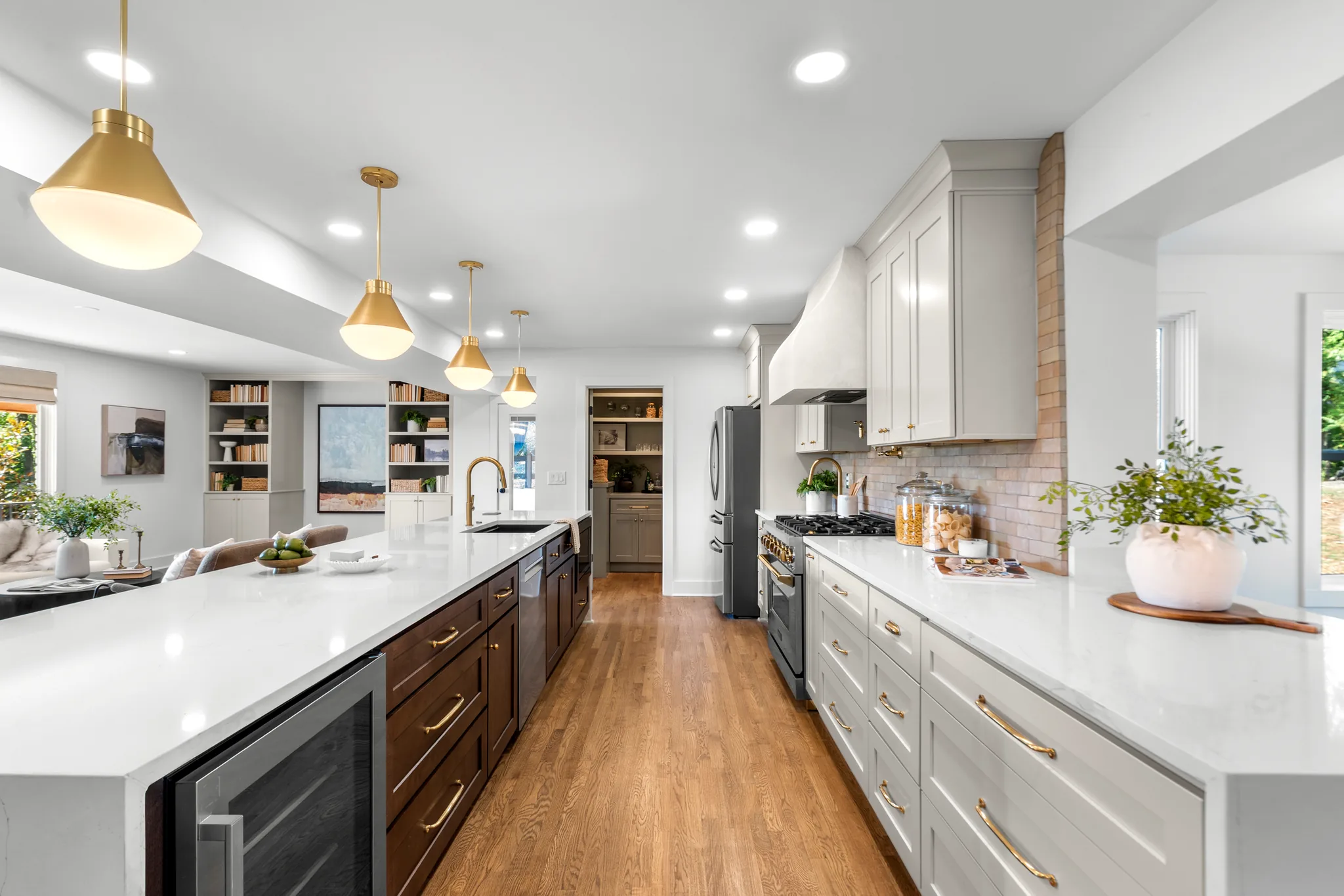 Open kitchen with white quartz counters, gray perimeter cabinets, walnut island, brass pendants and hardware, and oak floors leading to a pantry