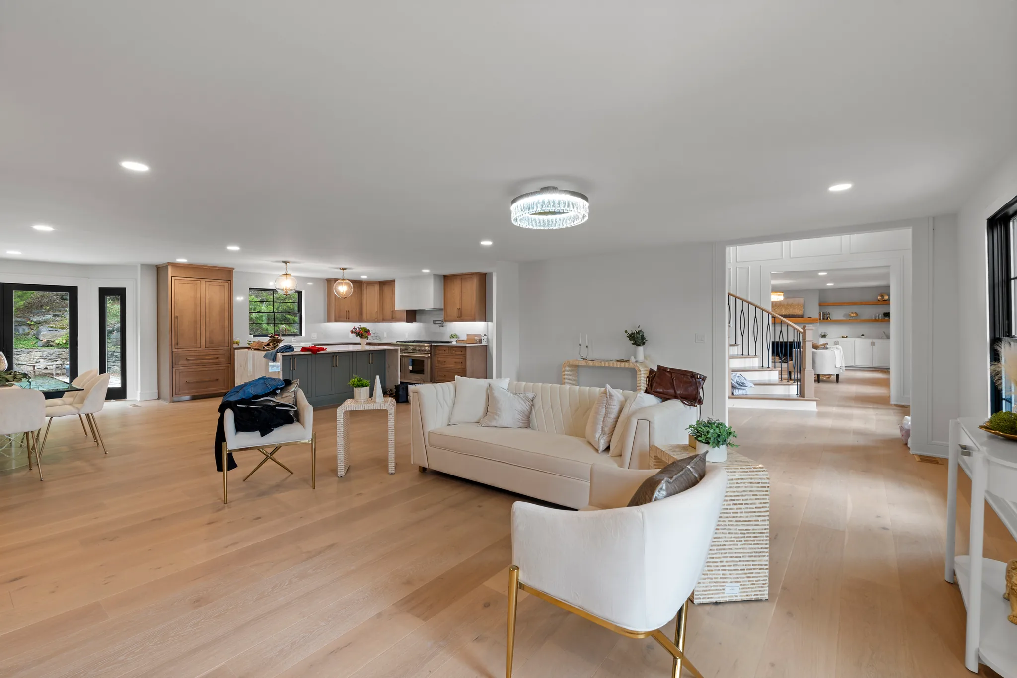 Open-plan living area with pale wide-plank wood floors, white sofa and chairs, a flush crystal ceiling light, and a wood-cabinet kitchen with gray island in the background