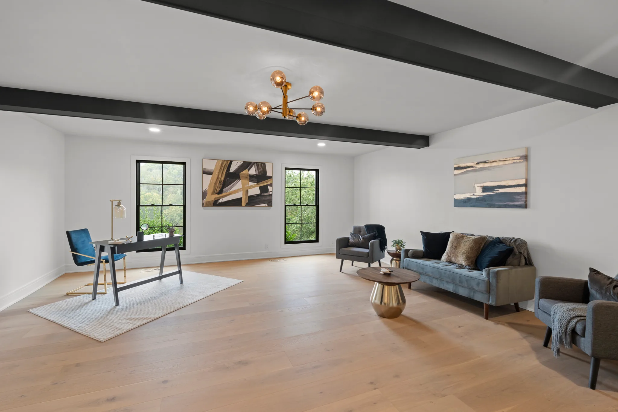 Open living and office space with white walls, exposed black ceiling beams, wide oak flooring, black-framed windows and a brass globe chandelier