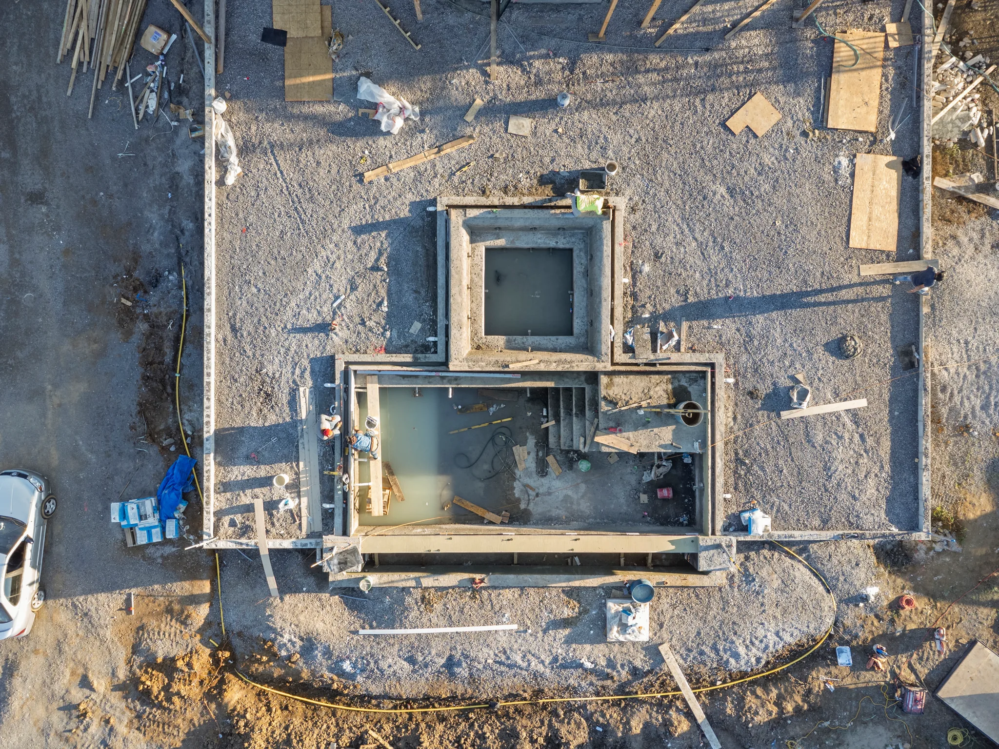 Aerial view of poured concrete foundation walls and footings on a gravel building pad, with lumber, forms and water pooled inside, under construction