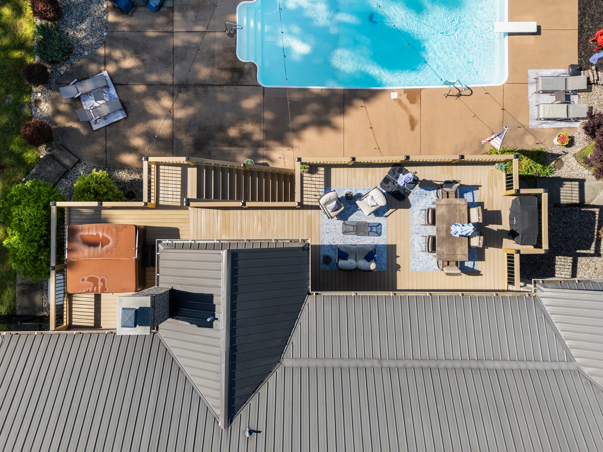 Aerial view looking straight down on a wood deck with pergola, hot tub, dining and lounge furniture beside a gray metal roof and concrete pool patio