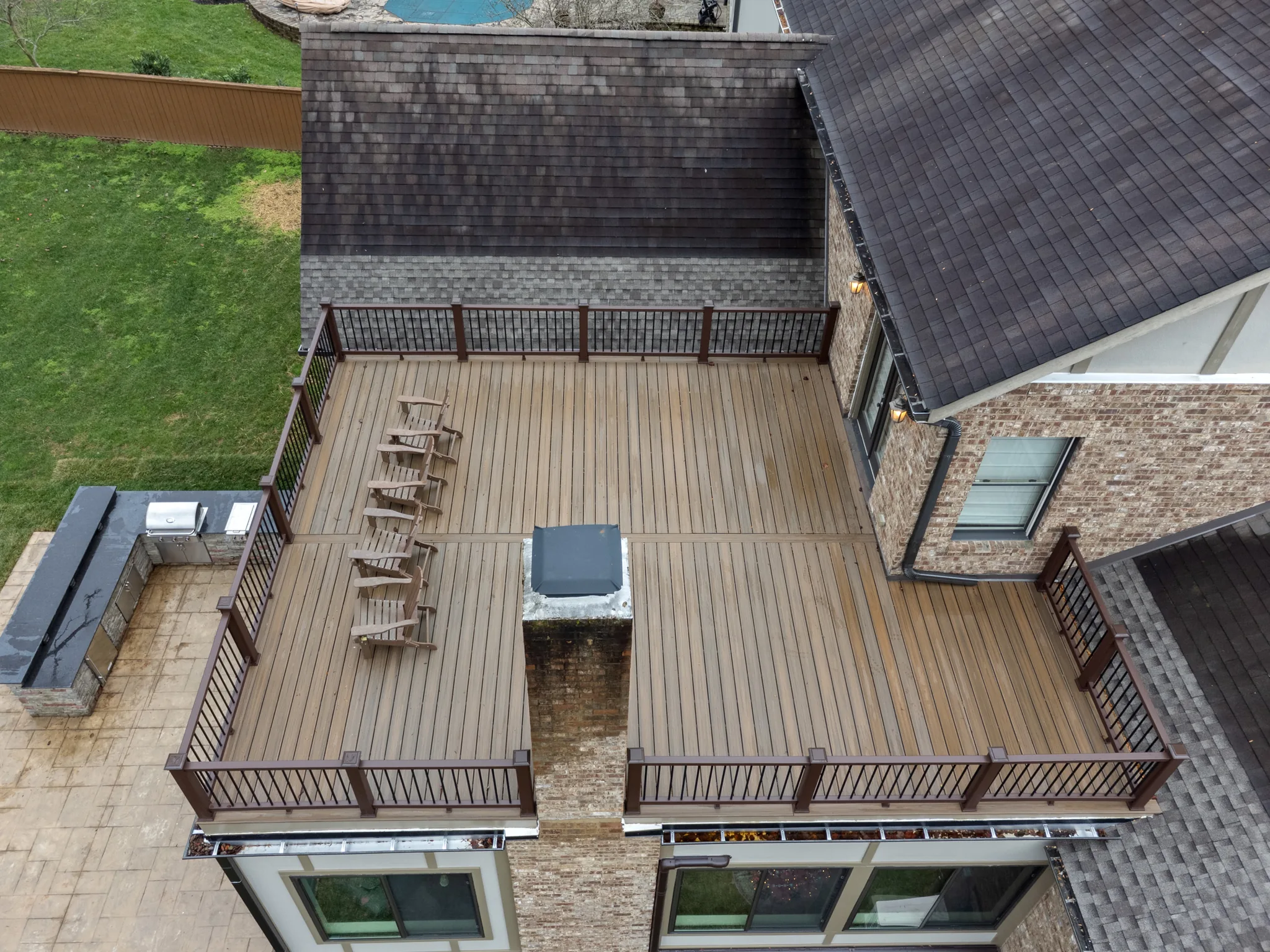 Aerial view of a raised composite deck with brown railings, a row of wood rocking chairs, brick chimney and adjacent stone outdoor kitchen on a paver patio below