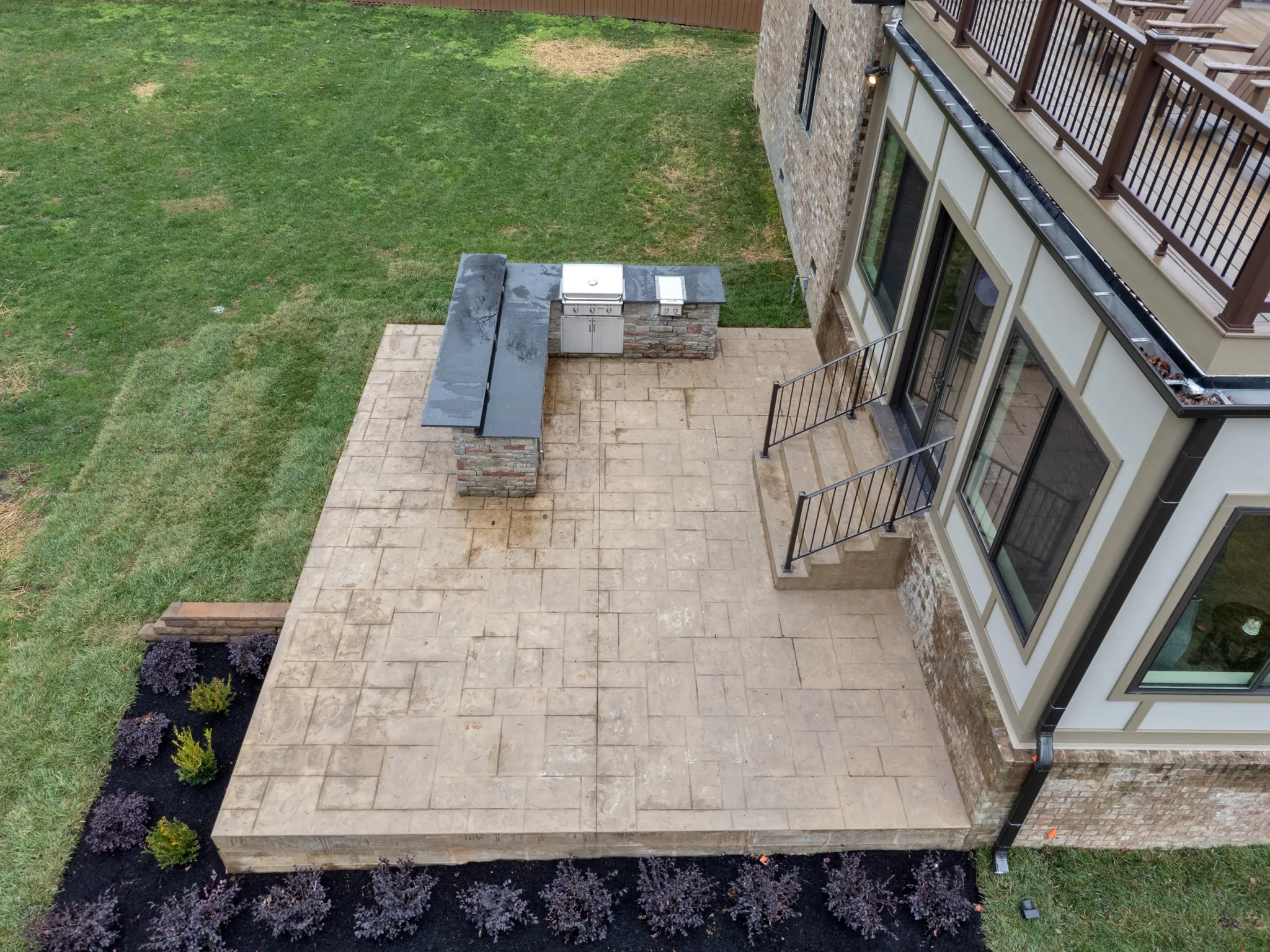 Aerial view of a stamped concrete patio with an L-shaped stone grill counter, dark stone tops, iron stair railing and new plantings