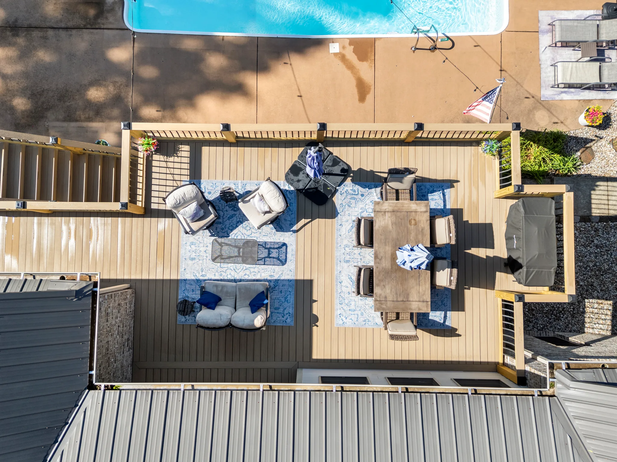 Aerial view looking straight down on a composite deck with wood pergola posts, patterned rugs, lounge chairs, dining table and covered hot tub beside a pool