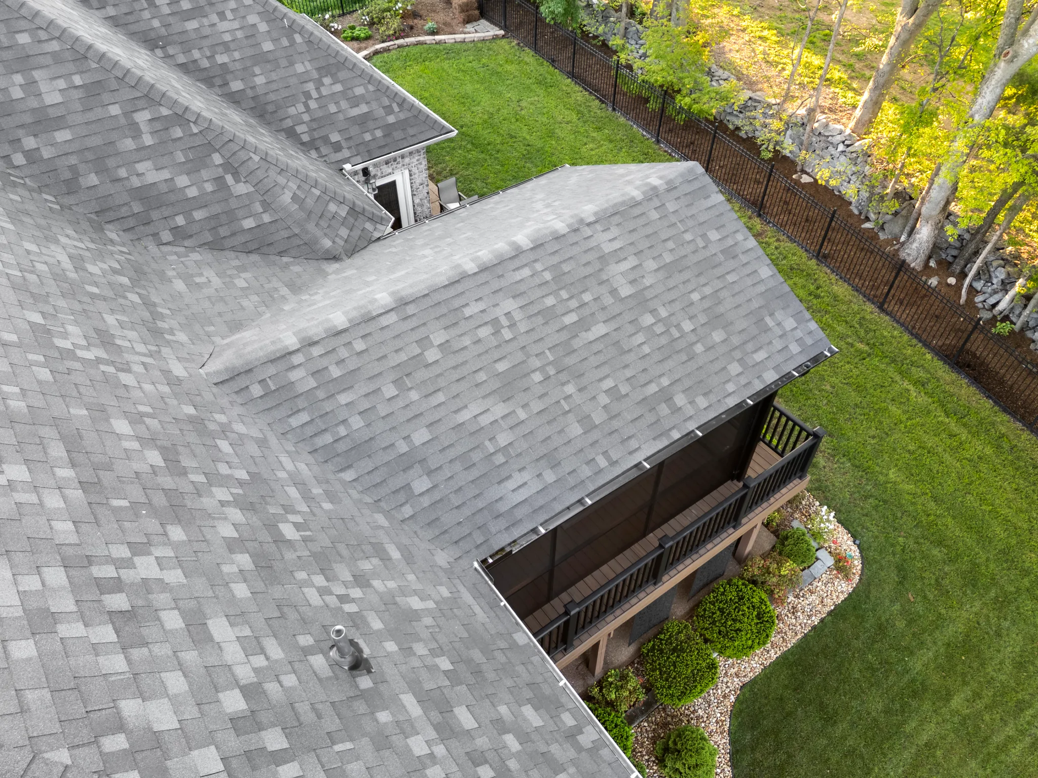 Aerial view of a gray shingled roof over a covered deck with black metal railing, wood columns and landscaped lawn beyond a black fence
