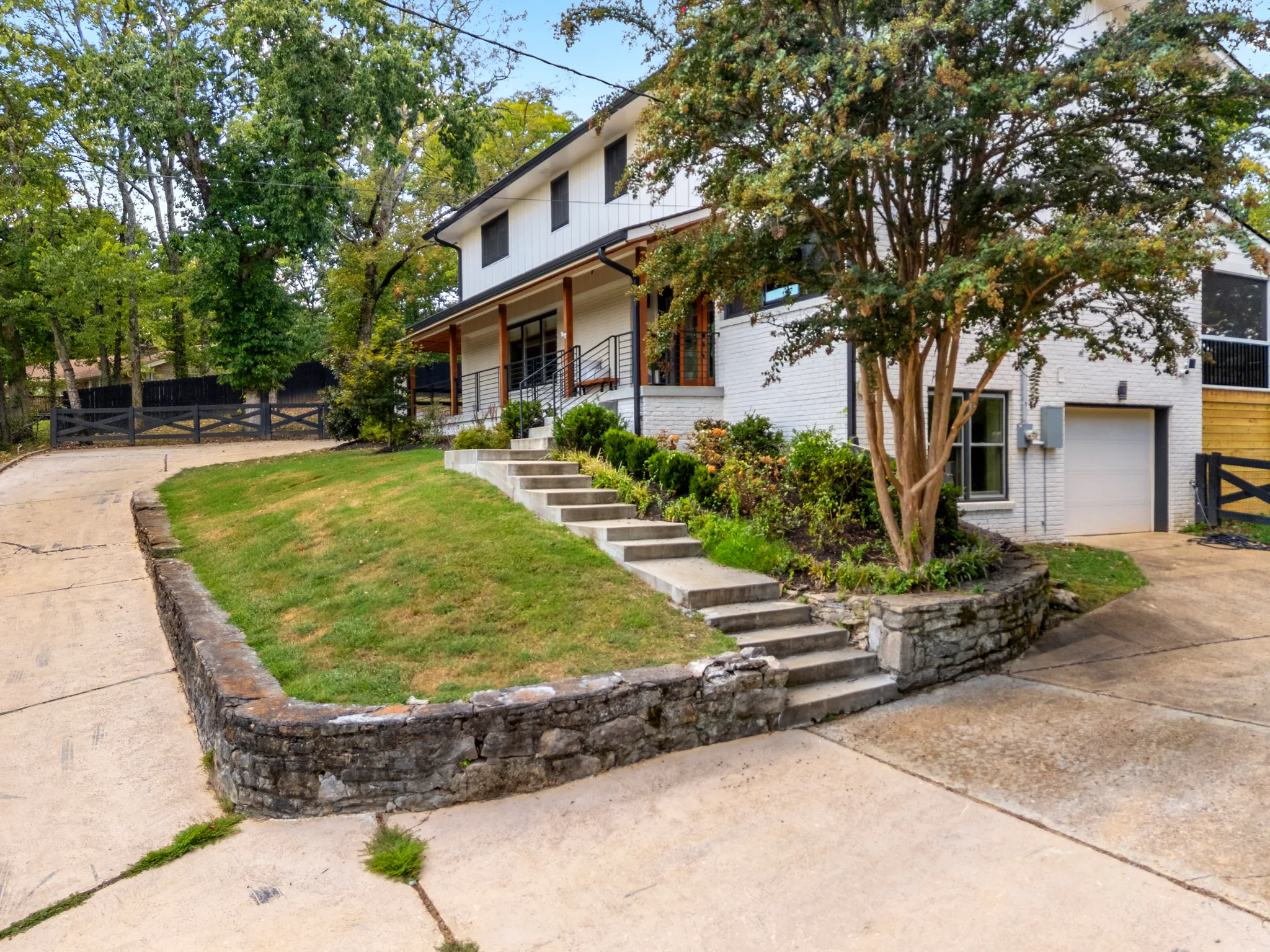 Two-story white painted brick and board and batten home behind a stacked stone retaining wall with concrete steps and a lawn