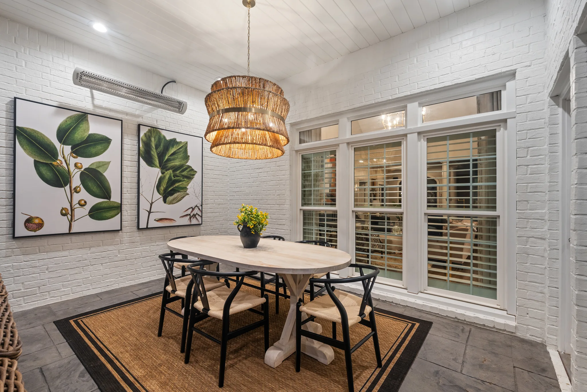 Enclosed porch with white painted brick walls, plank ceiling, flagstone floor, oval dining table, black chairs and a woven drum chandelier