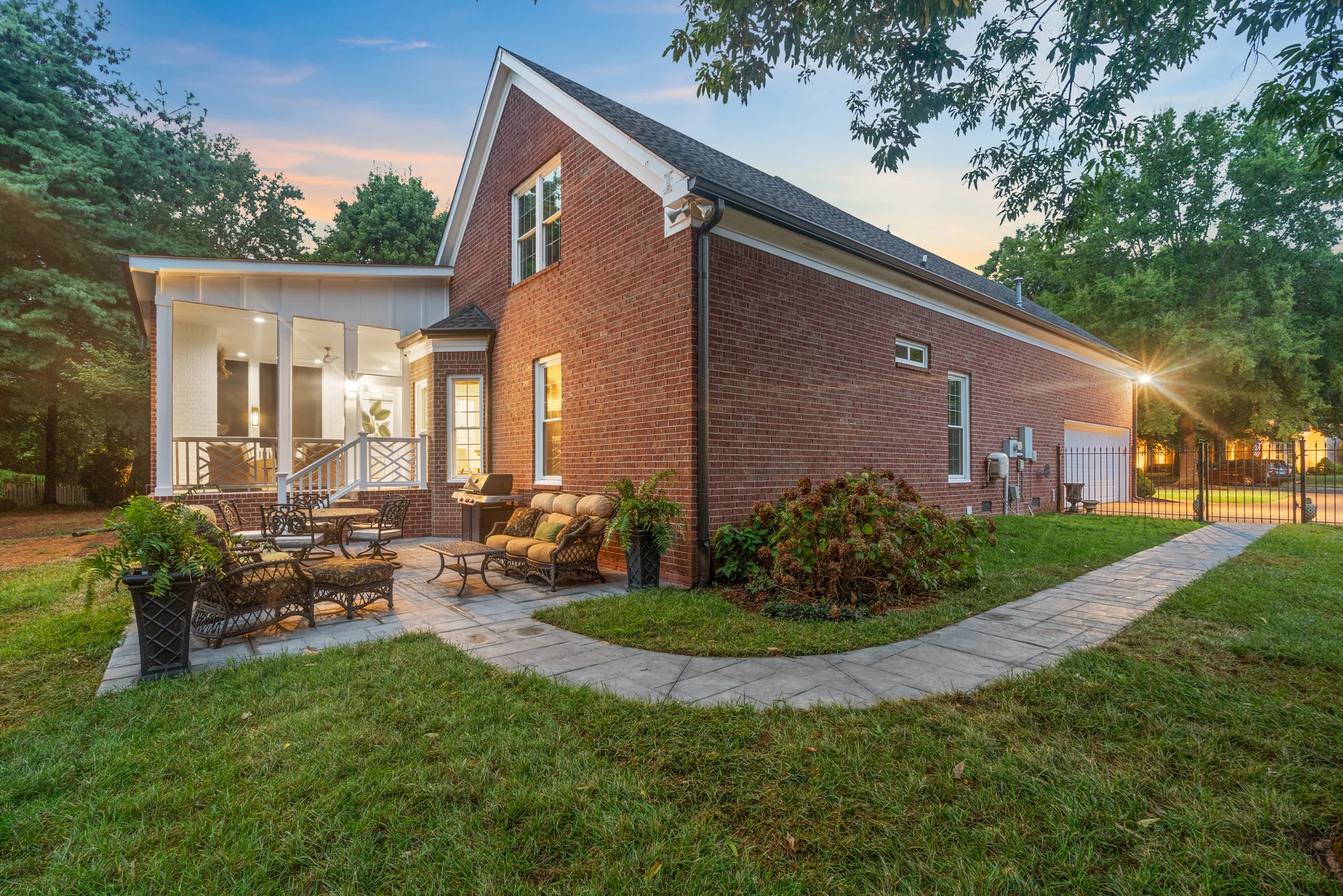 Paver patio with wicker lounge chairs and a grill beside a two-story red brick home with a screened porch at dusk