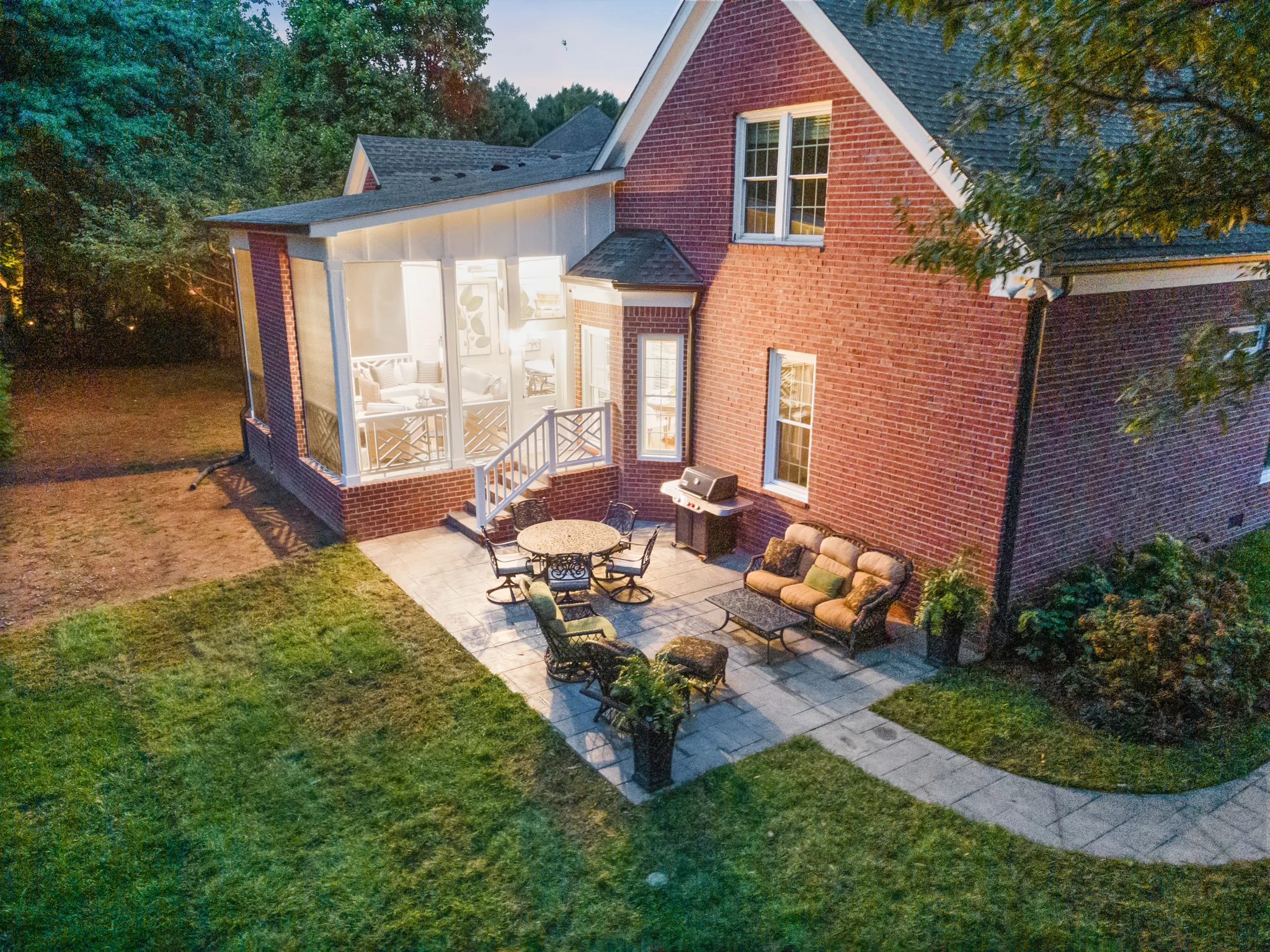 Aerial dusk view of a paver patio with cushioned seating and dining set beside a screened porch on a brick home