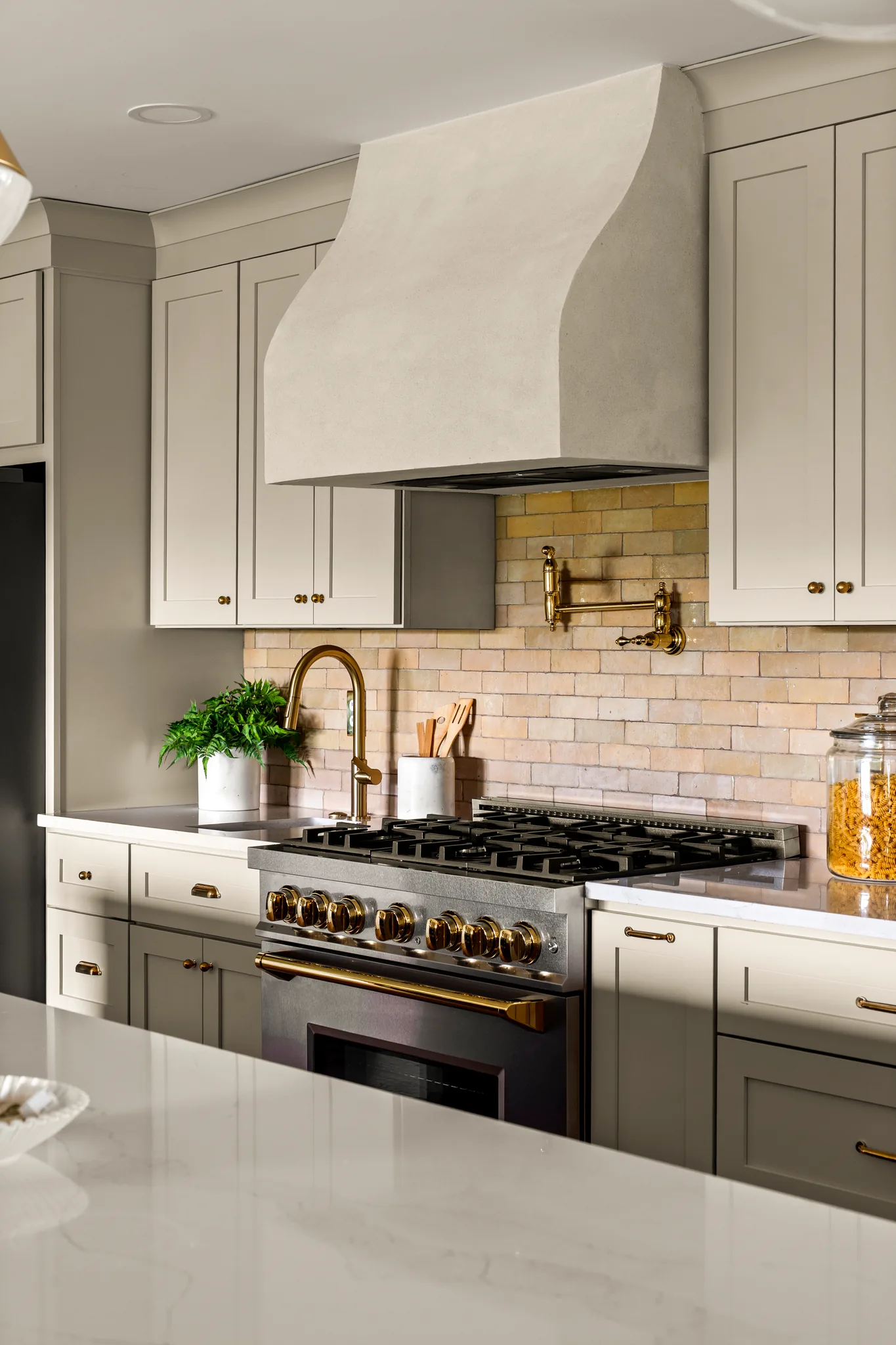 Kitchen range wall with a curved plaster hood, tan brick backsplash, brass pot filler and faucet, greige shaker cabinets and marble-look counters