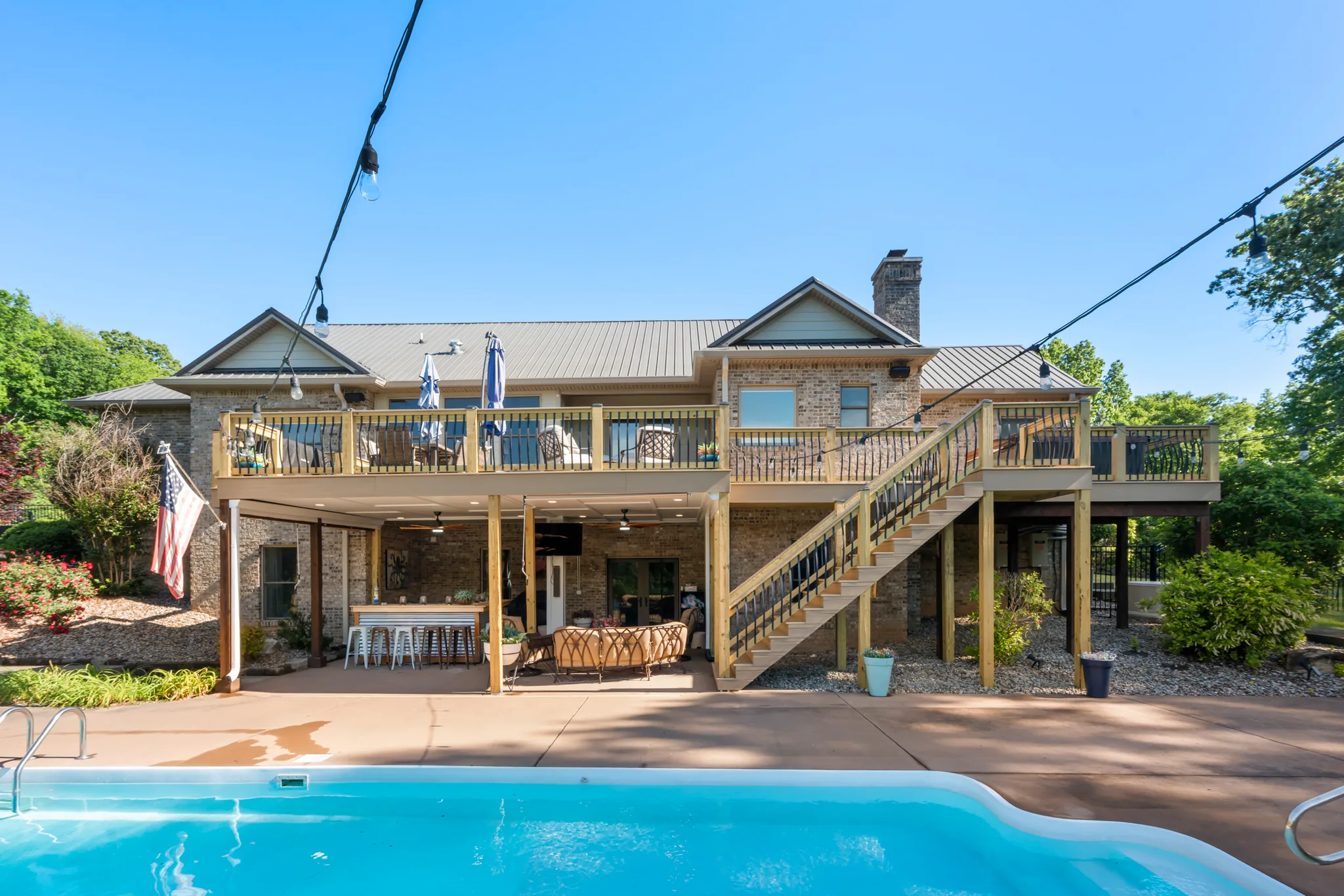 Rear of a brick home with a raised wood deck, covered patio bar and seating below, and an in-ground pool in the foreground