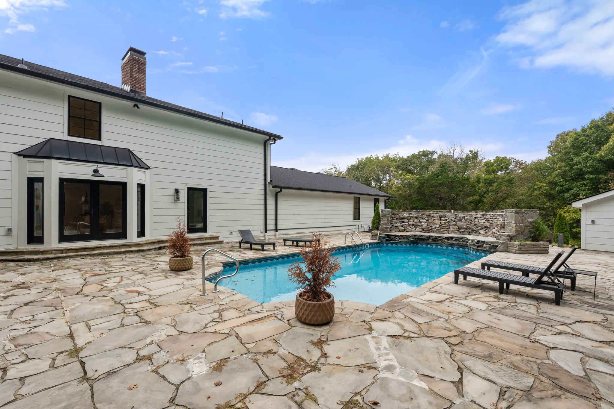 Rectangular pool surrounded by irregular flagstone paving behind a white home with black windows, brick chimney and a dry-stacked stone retaining wall