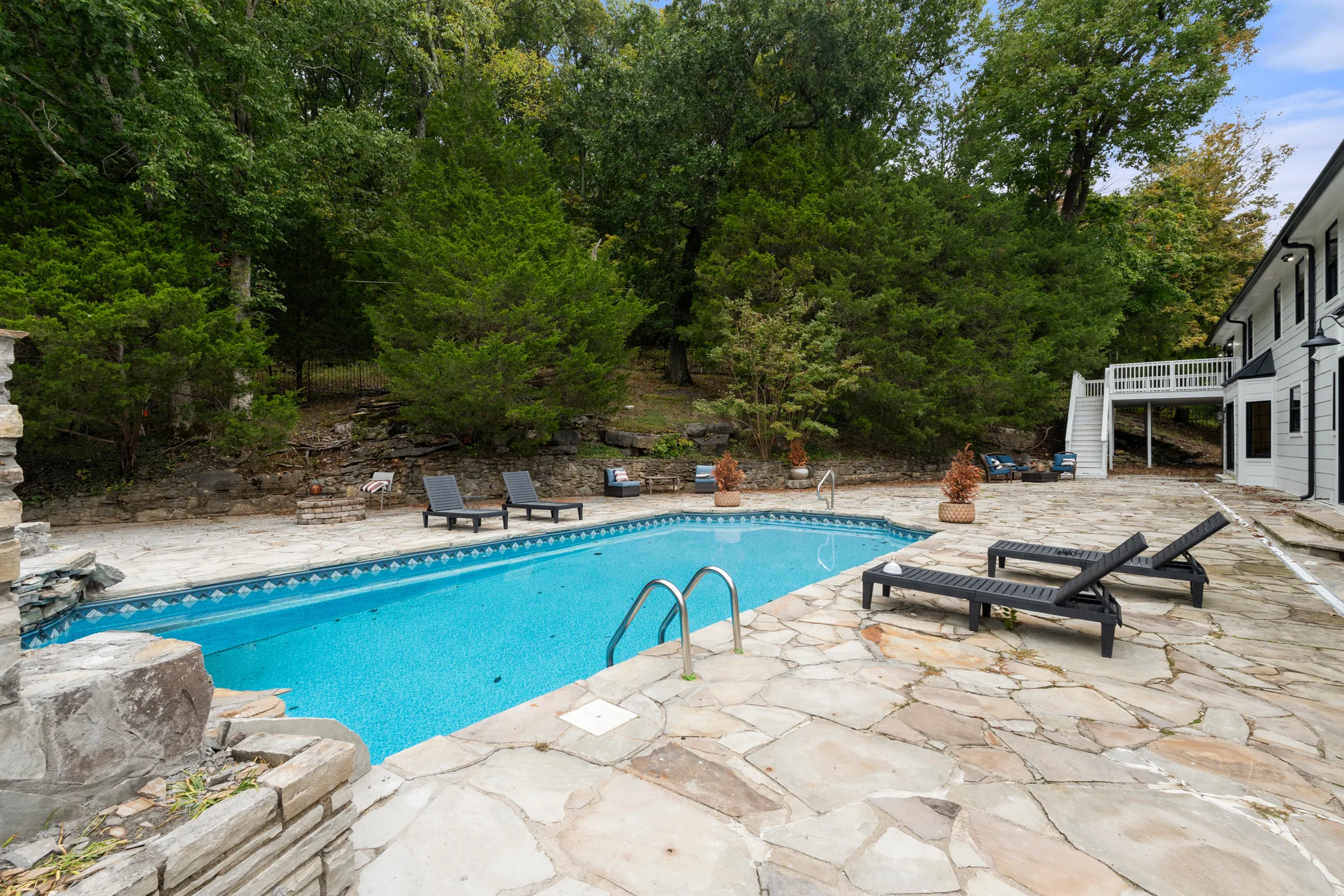 Rectangular pool with blue tile border set in an irregular flagstone patio, black loungers alongside and a wooded hillside with stone retaining wall behind