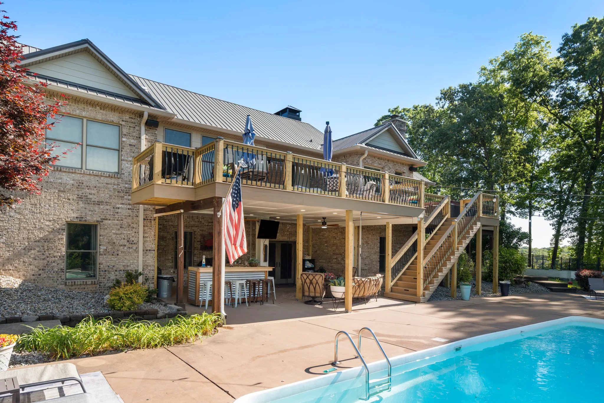 Brick home with an upper wood deck sheltering a covered poolside bar with metal-front counter, stools and ceiling fans beside a concrete pool patio