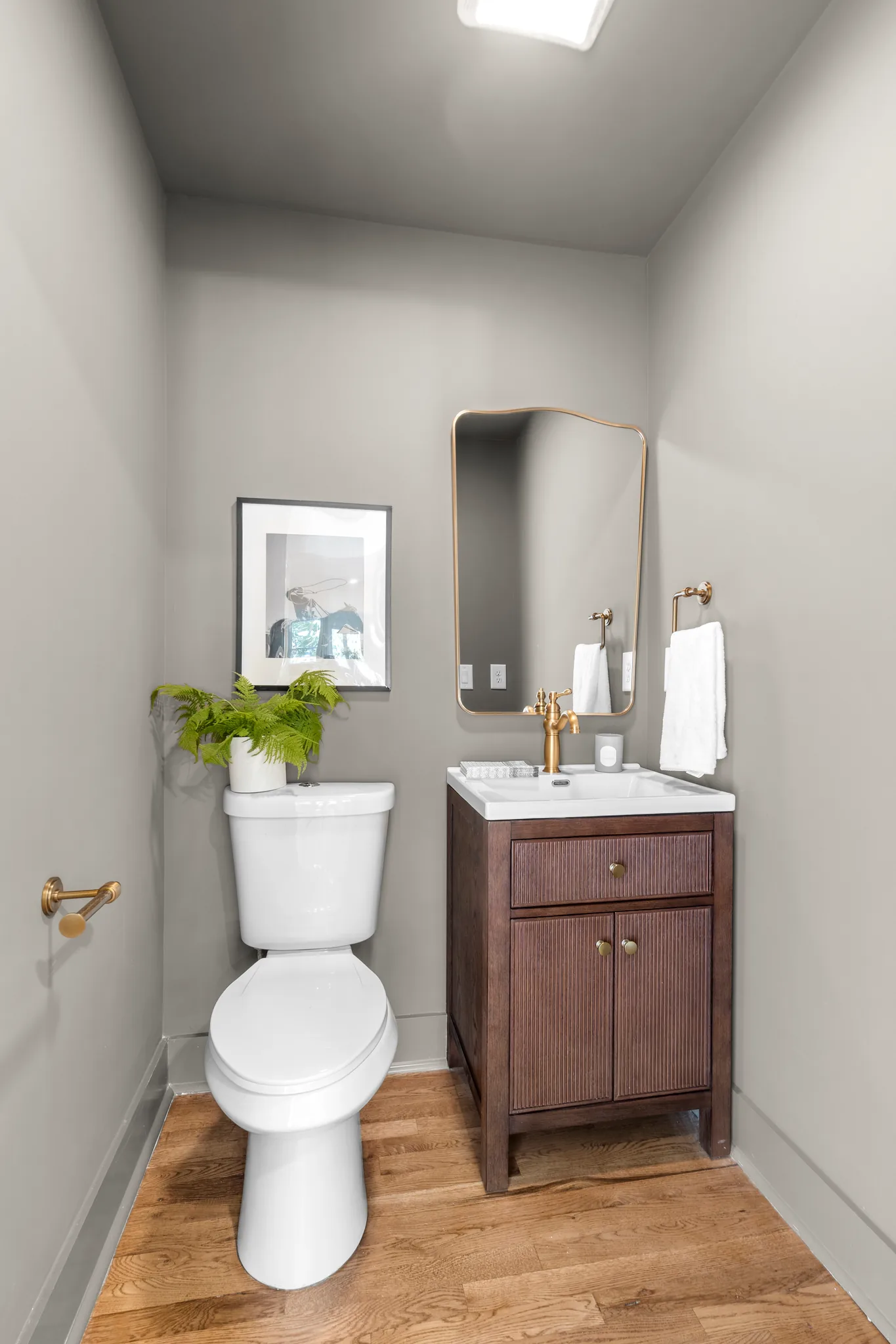 Small powder room with a dark fluted wood vanity, brass faucet, gold-framed mirror, gray walls, white toilet and oak floors